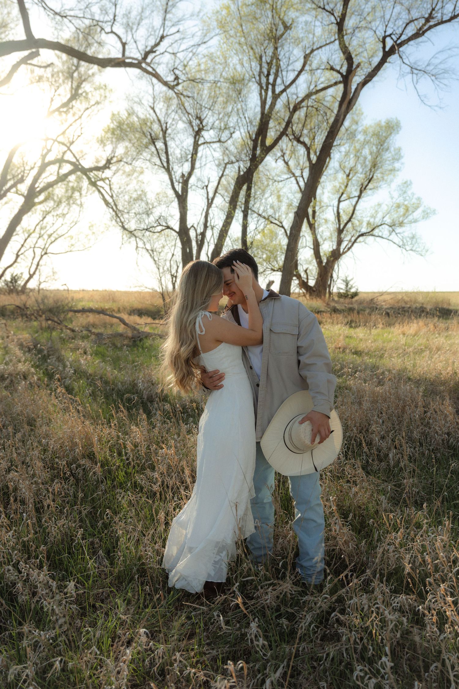 Couple sharing a romantic kiss in a sunlit field, bride in white dress, groom holding cowboy hat, trees in background.