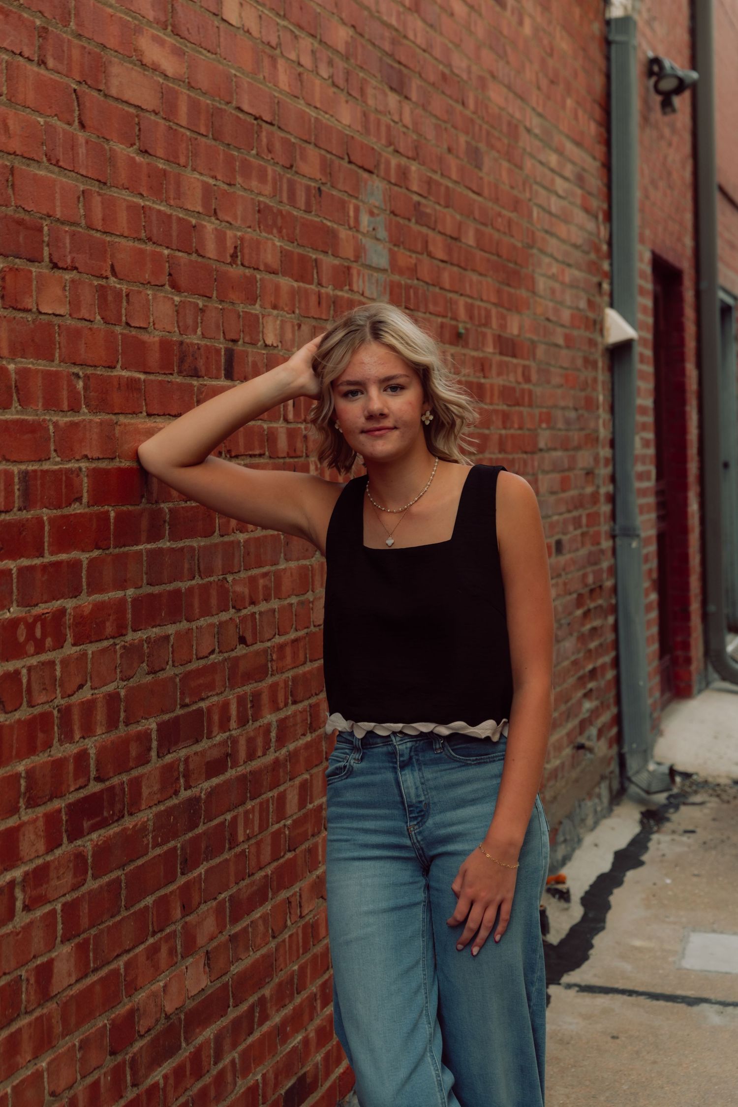 Woman in black tank top and jeans poses against red brick wall in urban setting.