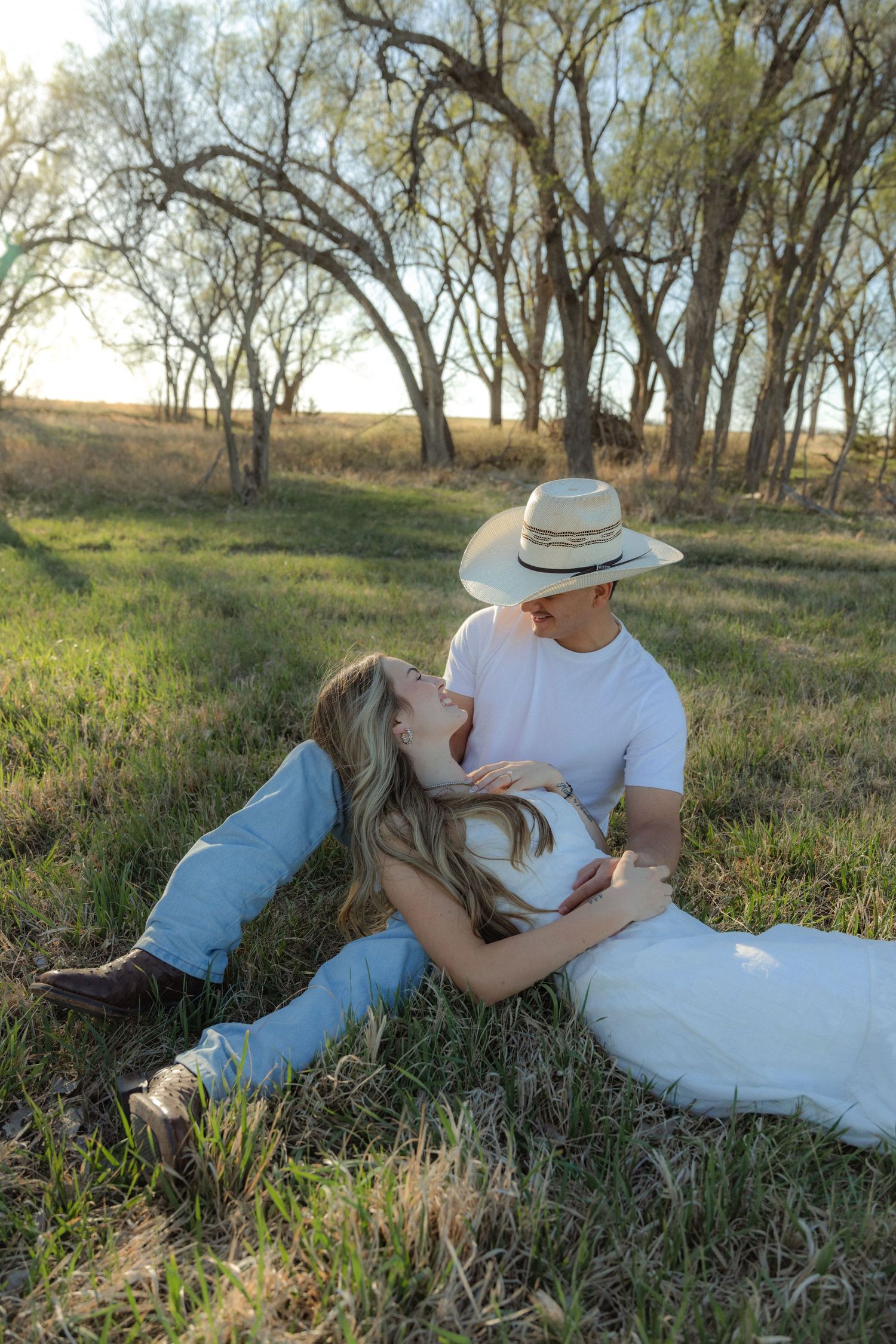 A couple shares a romantic moment in a grassy field, with the man in a cowboy hat gazing down at the woman lying in his arms.
