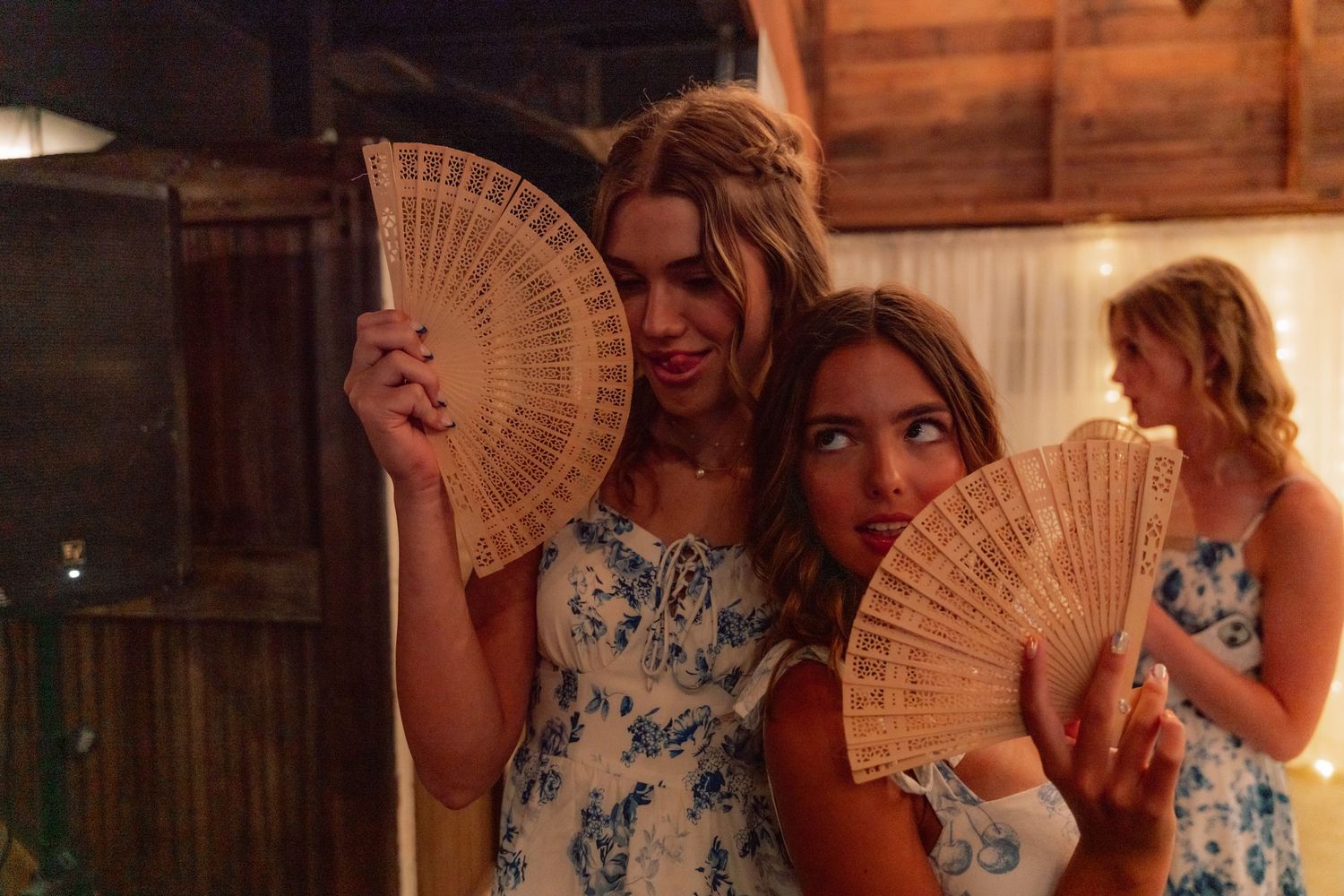 Bridesmaids in matching floral dresses hold decorative fans at a rustic wedding venue.
