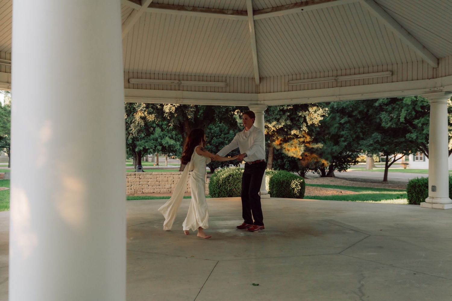 A couple in formal attire dances in an elegant outdoor pavilion surrounded by gardens.
