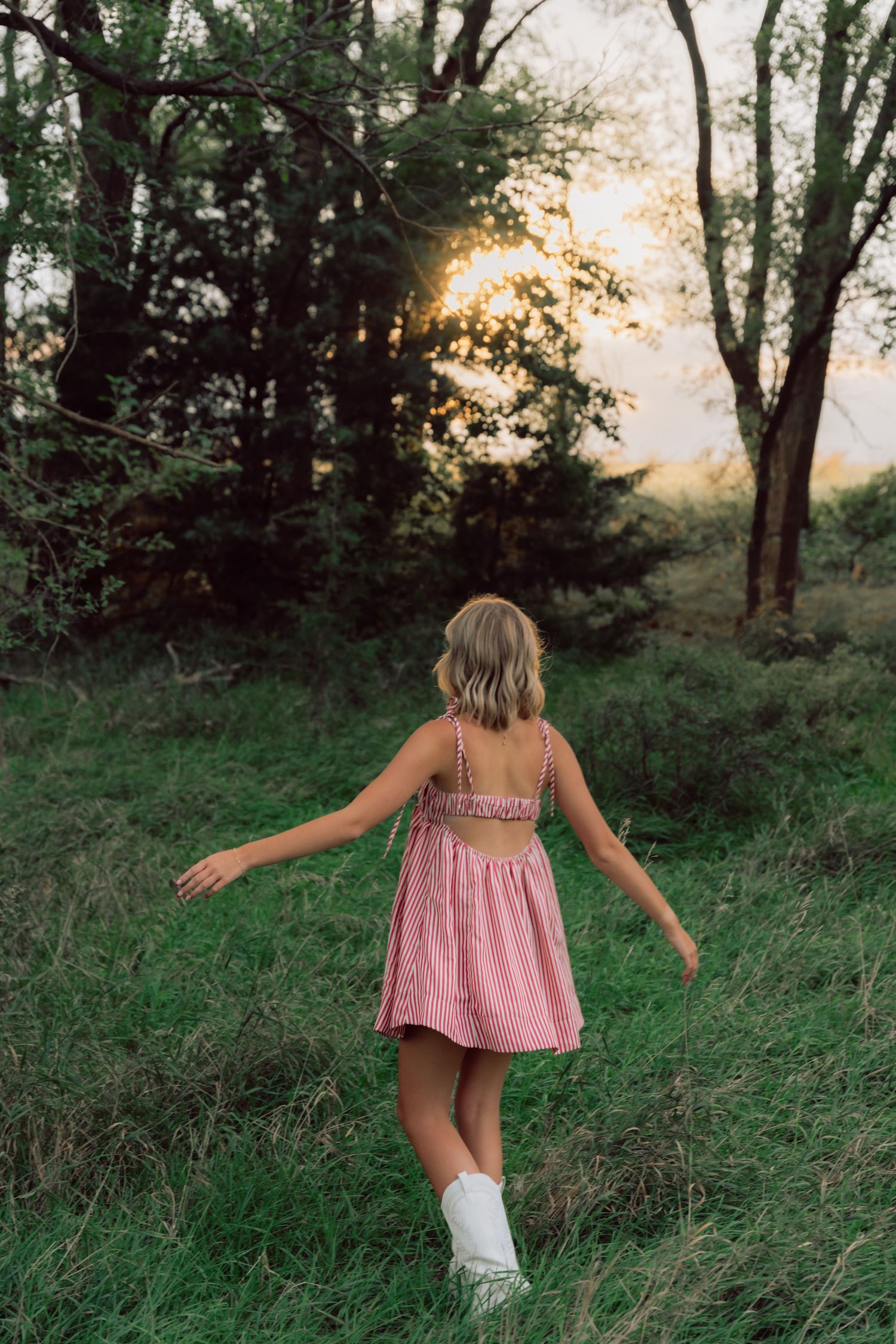 Person in pink dress walking through tall grass at sunset with arms outstretched.