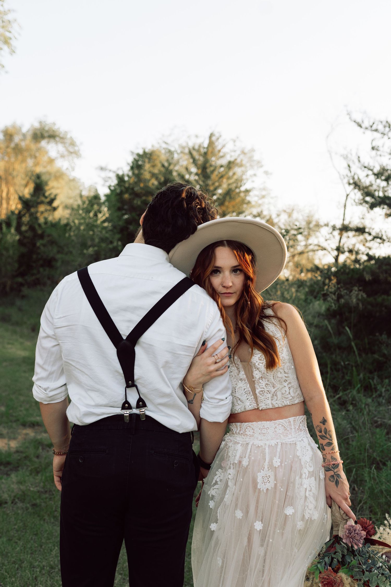 A bride in a sheer lace dress and groom in suspenders pose together in a natural outdoor setting.