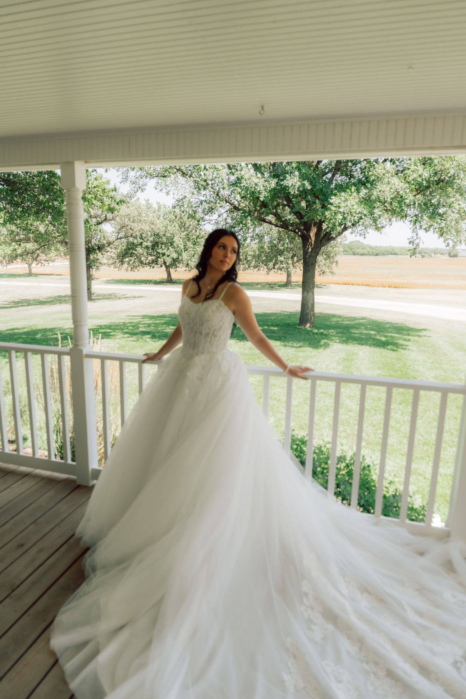 A bride in a flowing white ballgown twirls on a covered porch with trees visible in the background.