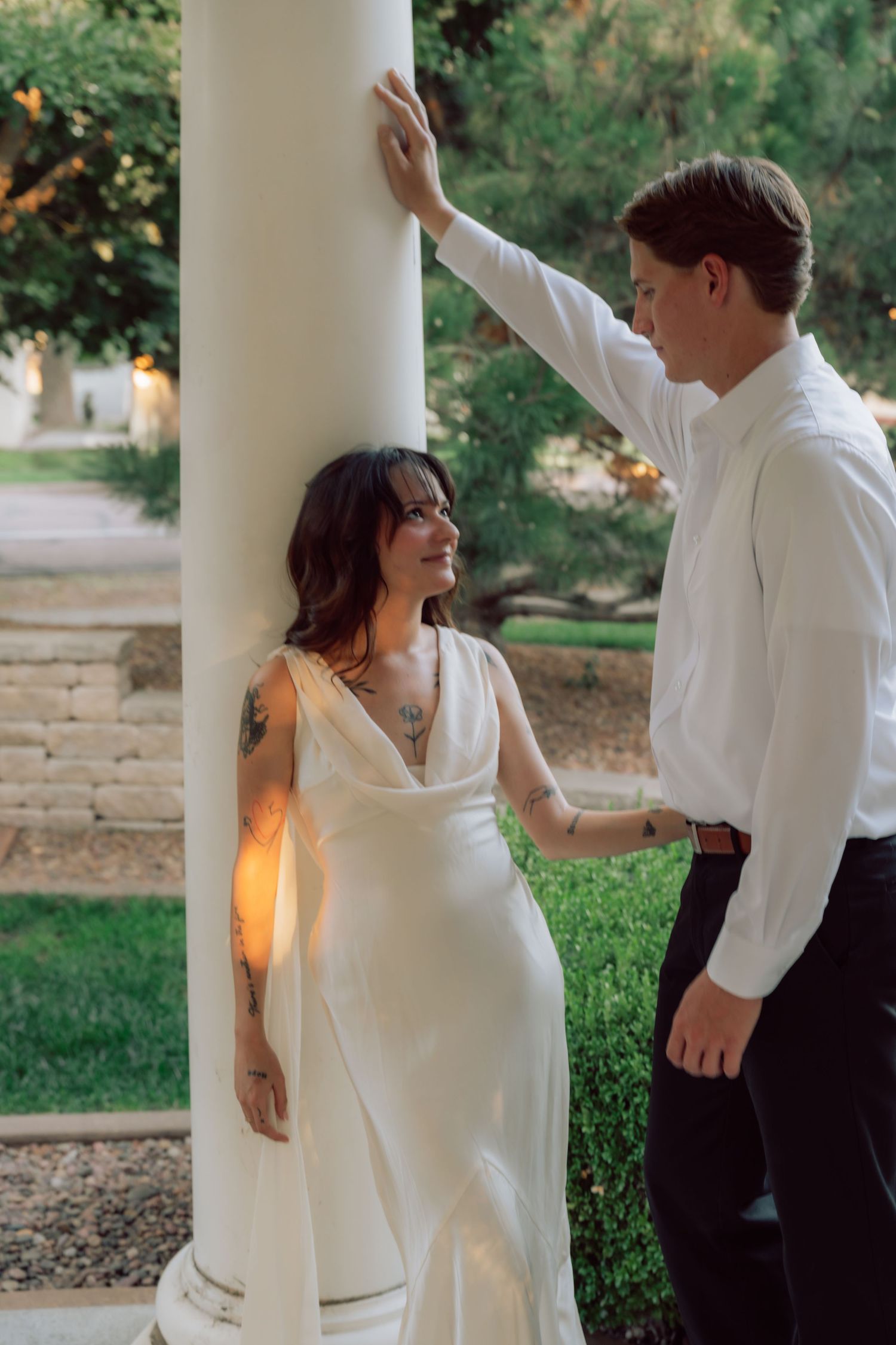 A couple in white clothing poses romantically against a classical column in a garden setting.