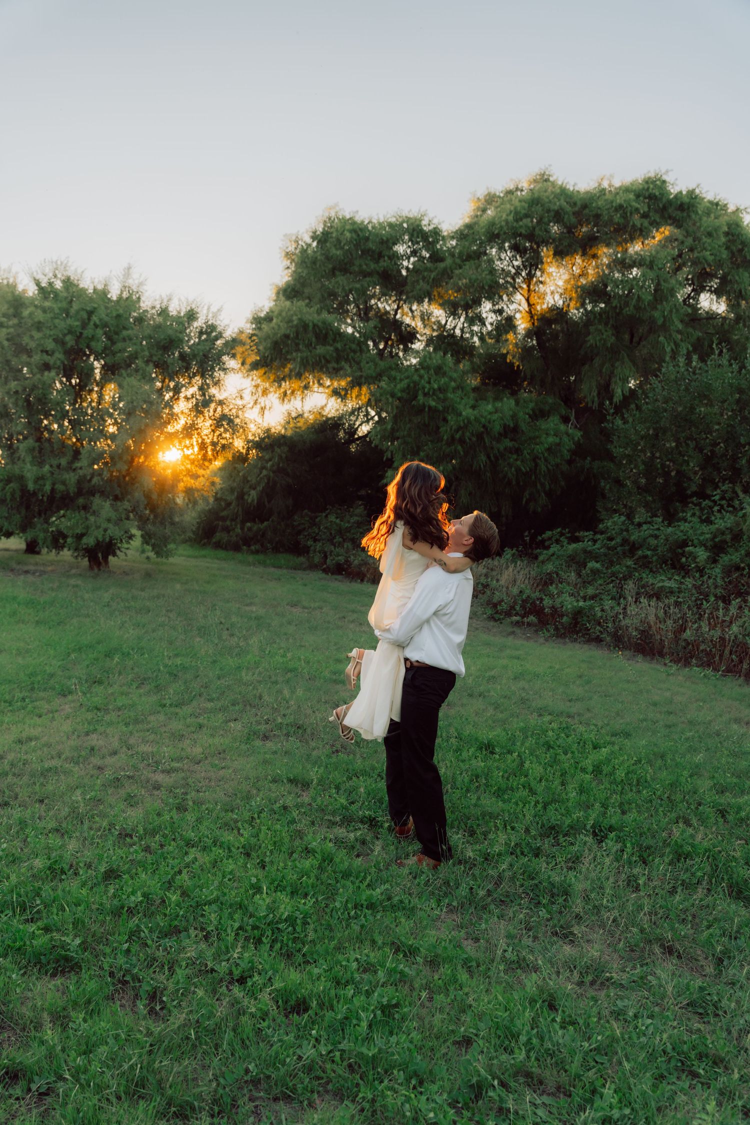 A couple dances playfully in a grassy field during golden hour with trees in the background.