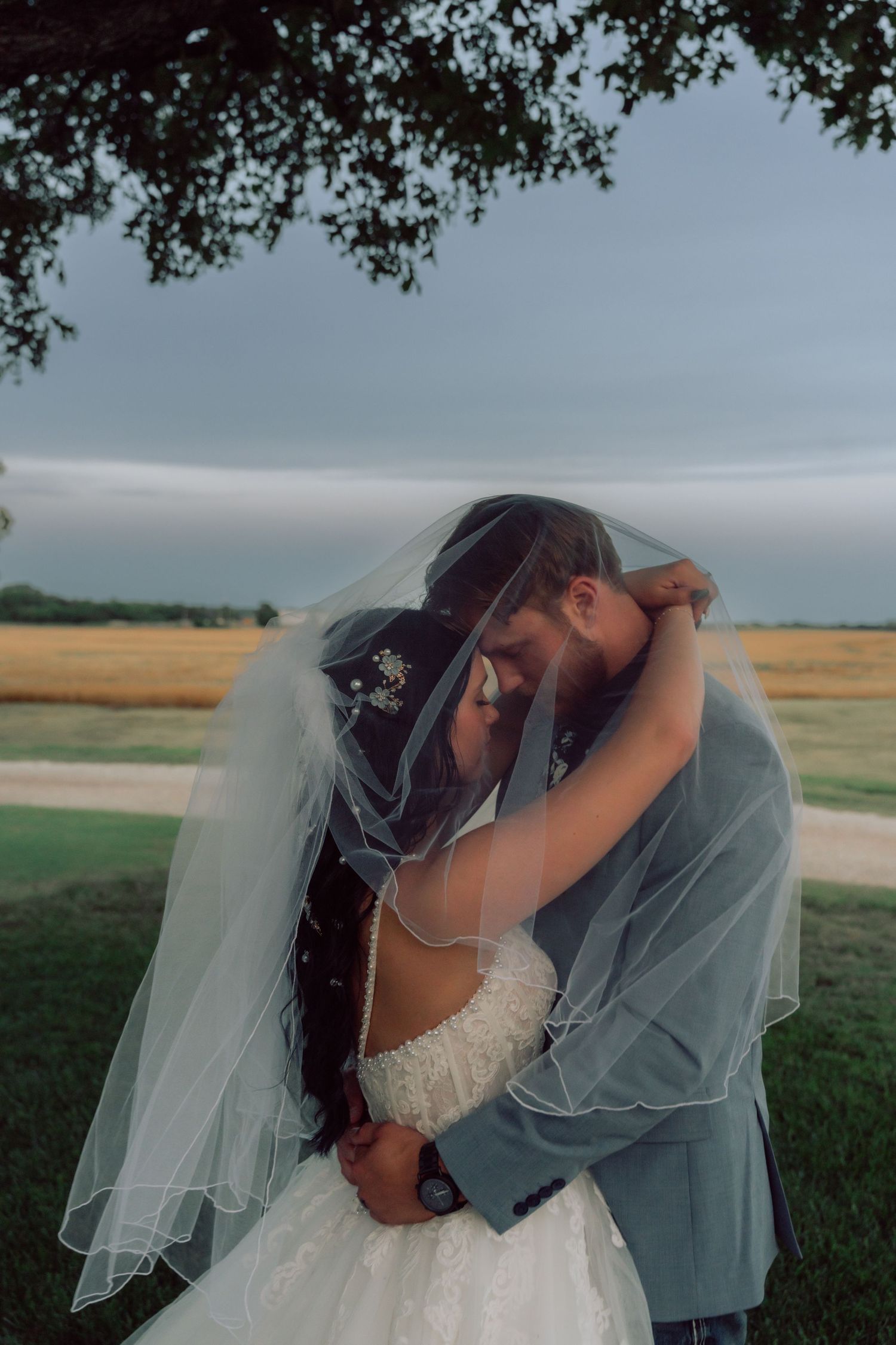 A romantic wedding photo showing a couple embracing under tree branches in an outdoor field setting.