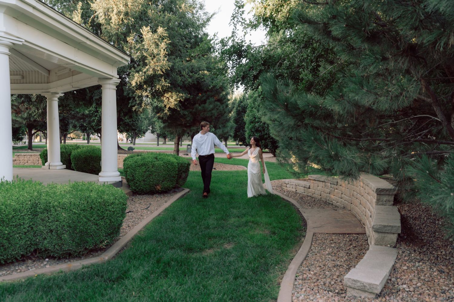 A couple walks along a curved garden path next to a white pavilion structure.