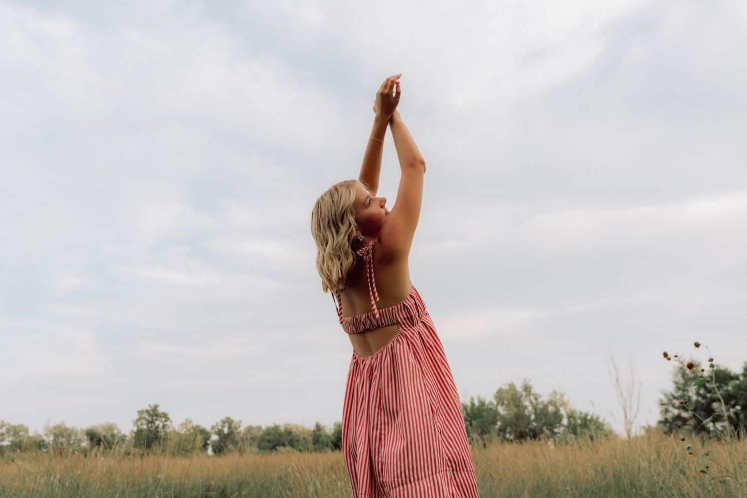 Person in pink dress reaches towards sky in open field during sunset.