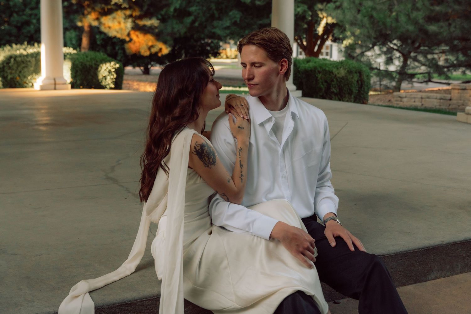 A couple in white attire shares a tender moment under a covered pavilion at dusk.