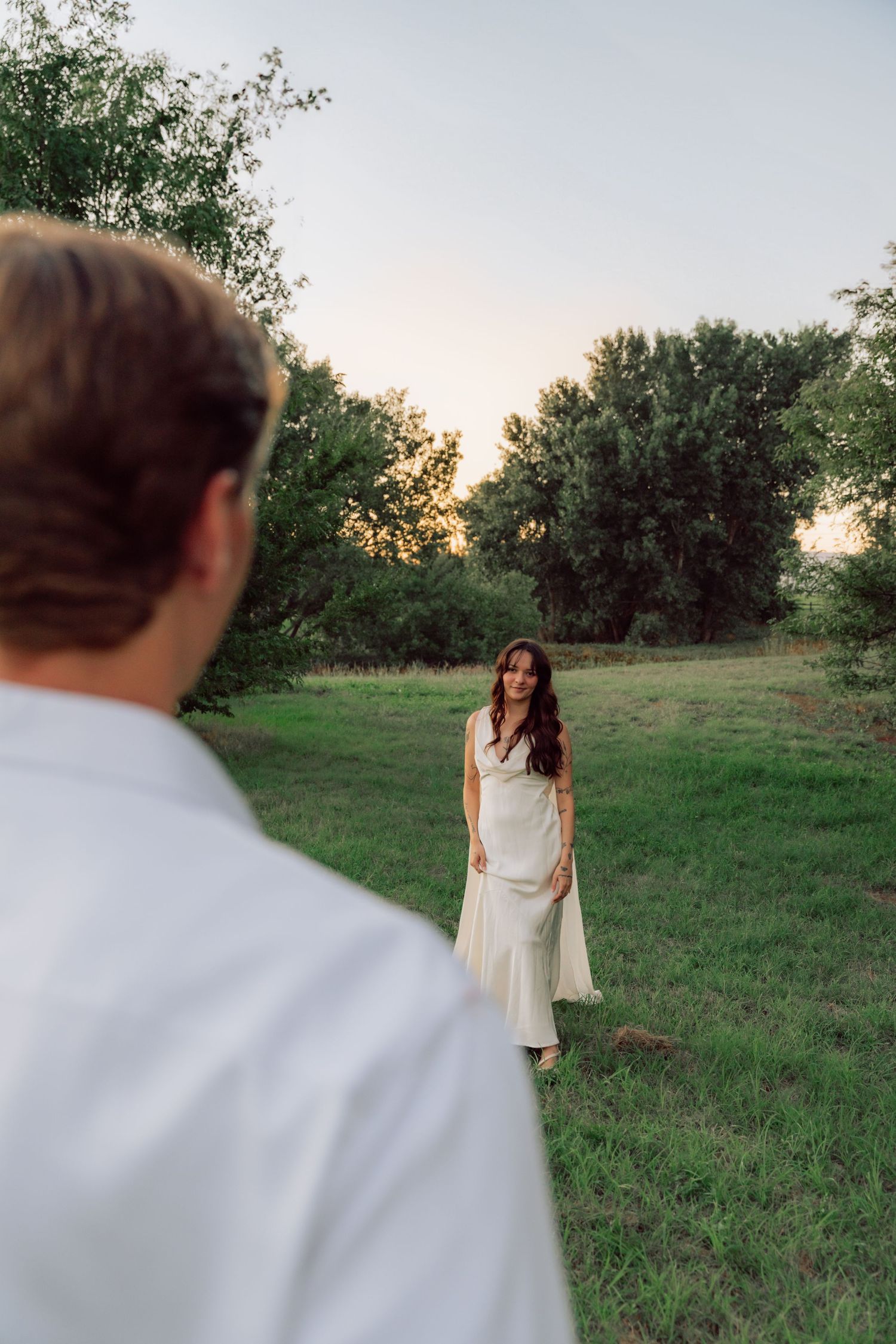 Two people in formal wear standing apart in a grassy field at sunset.