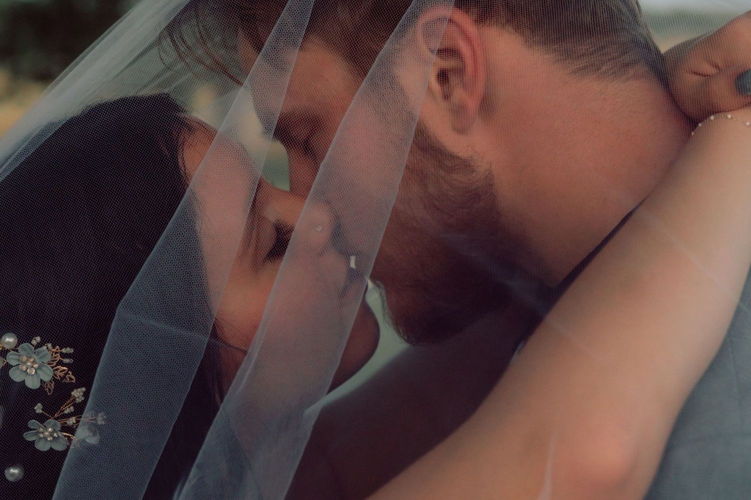 Newlyweds share a tender kiss under a flowing white veil against a natural outdoor backdrop.