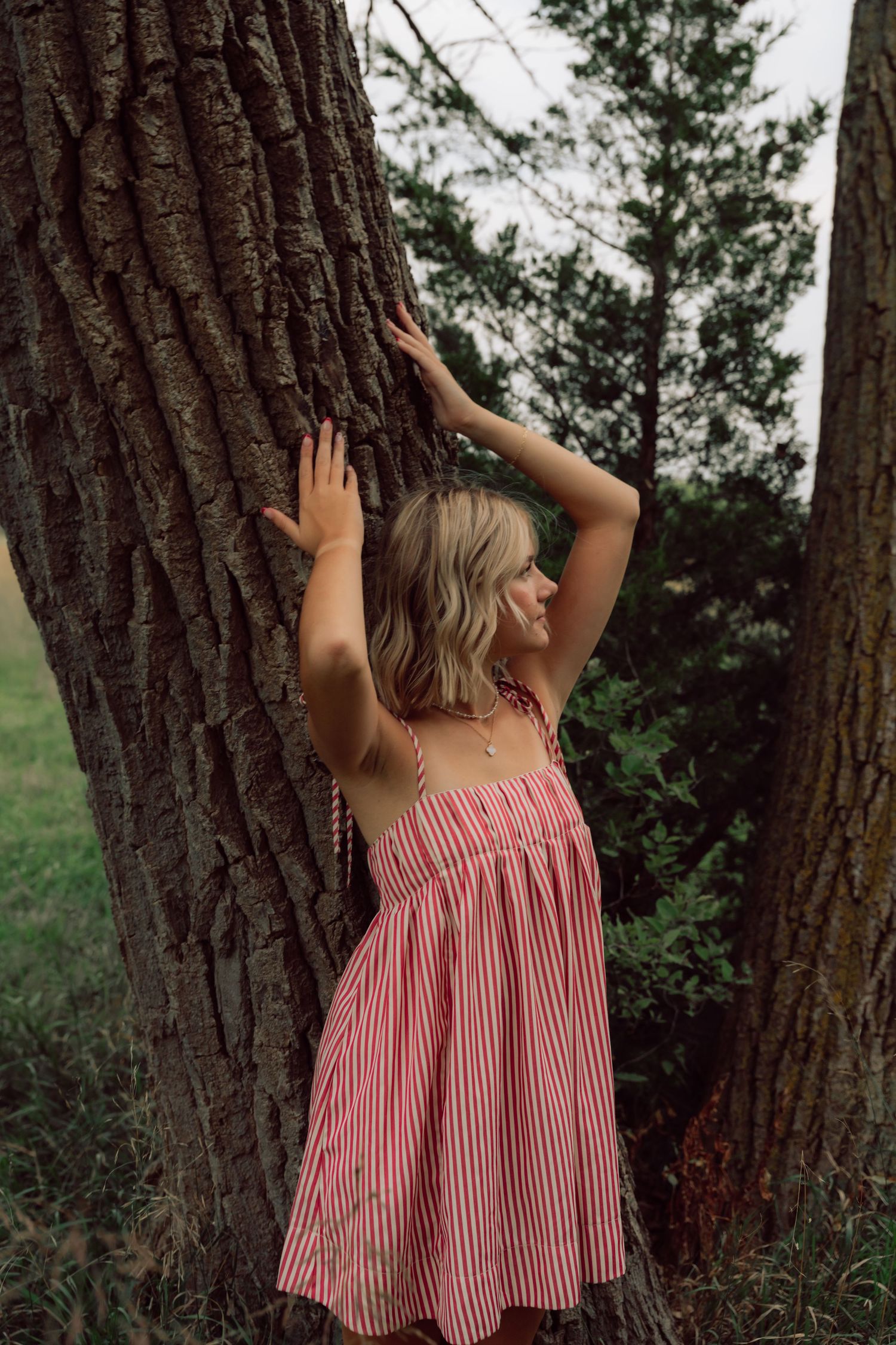 Person in striped pink dress poses against large tree trunk in natural setting.