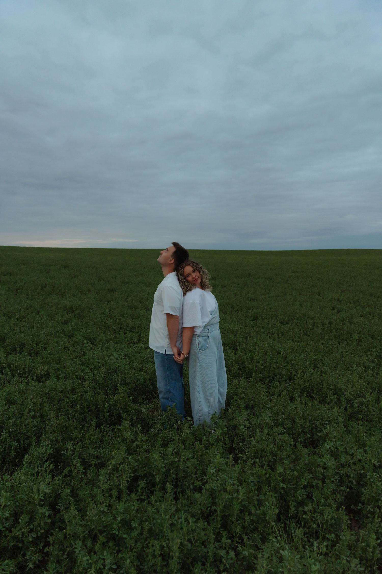 Couple standing back-to-back in a lush green field under a cloudy sky, sharing a tender romantic moment outdoors.