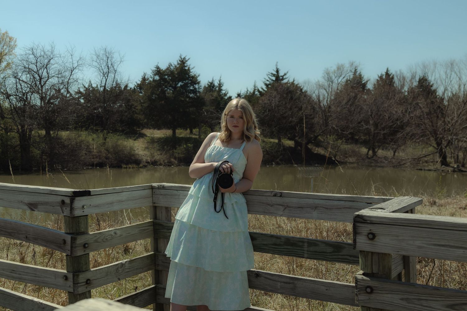 Blonde woman in white tiered dress on wooden bridge overlooking pond and trees on a sunny day.