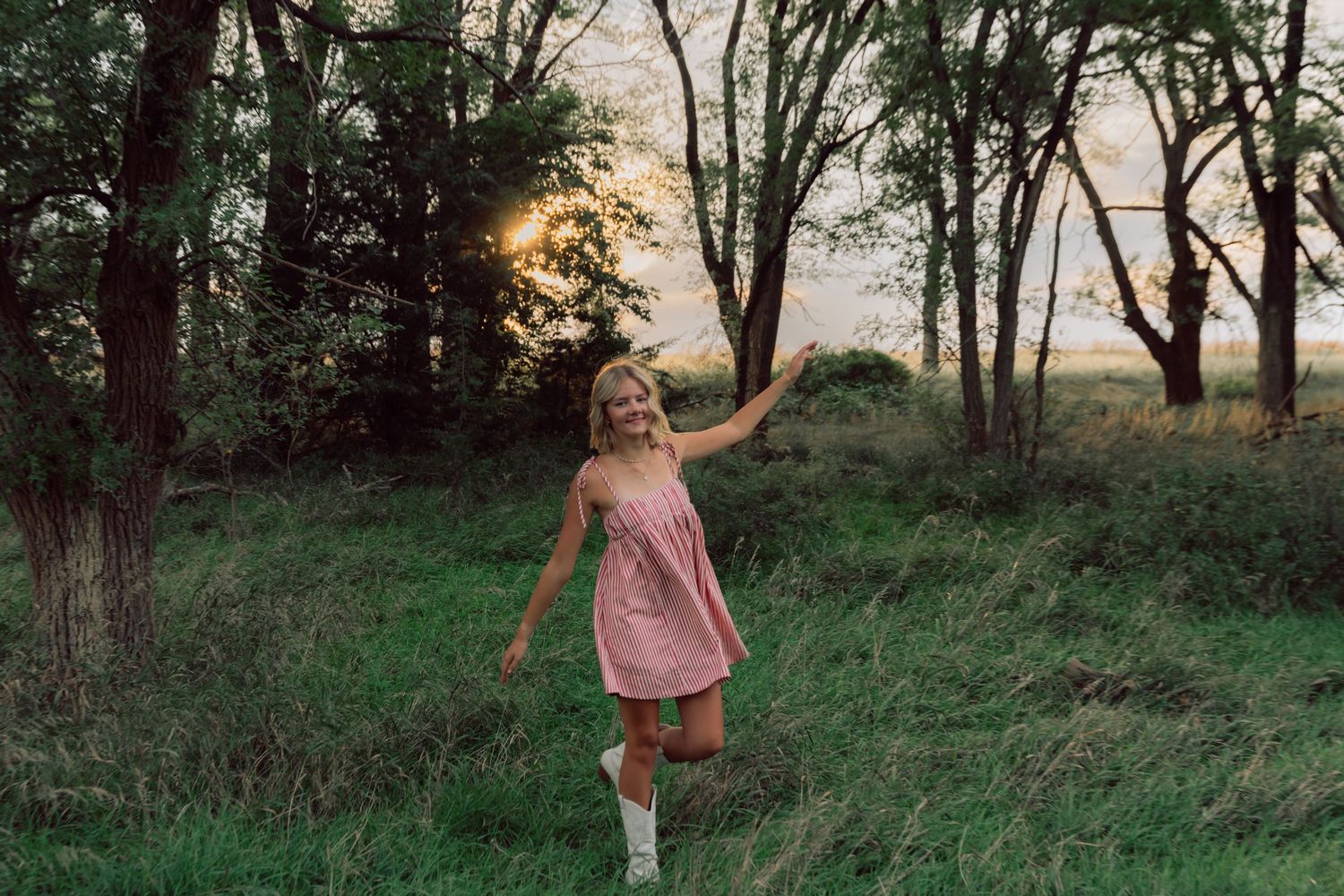 Person in pink sundress dancing in grassy field surrounded by trees at sunset.