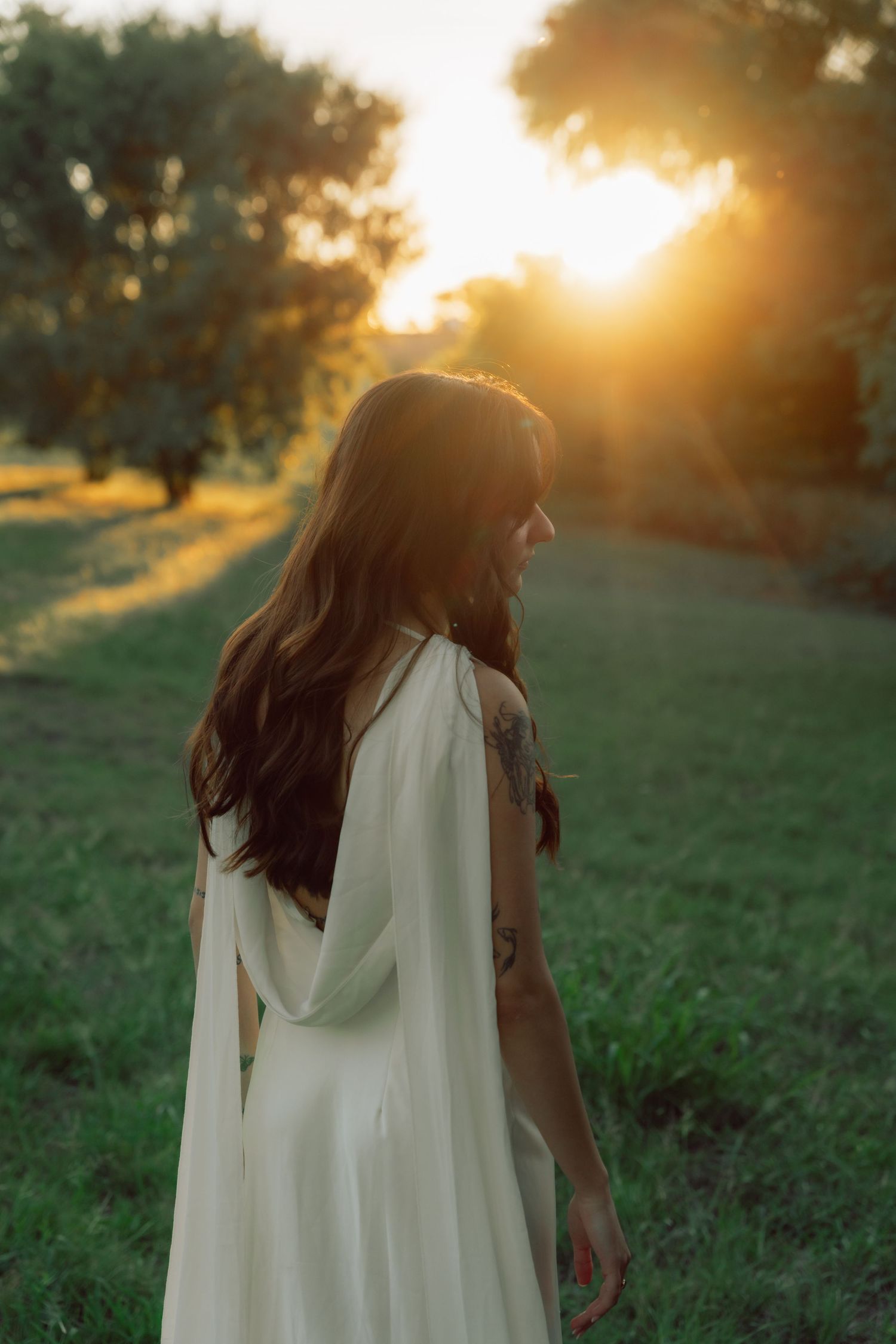 A figure in a flowing white dress stands in a grassy field at sunset with golden light filtering through trees.