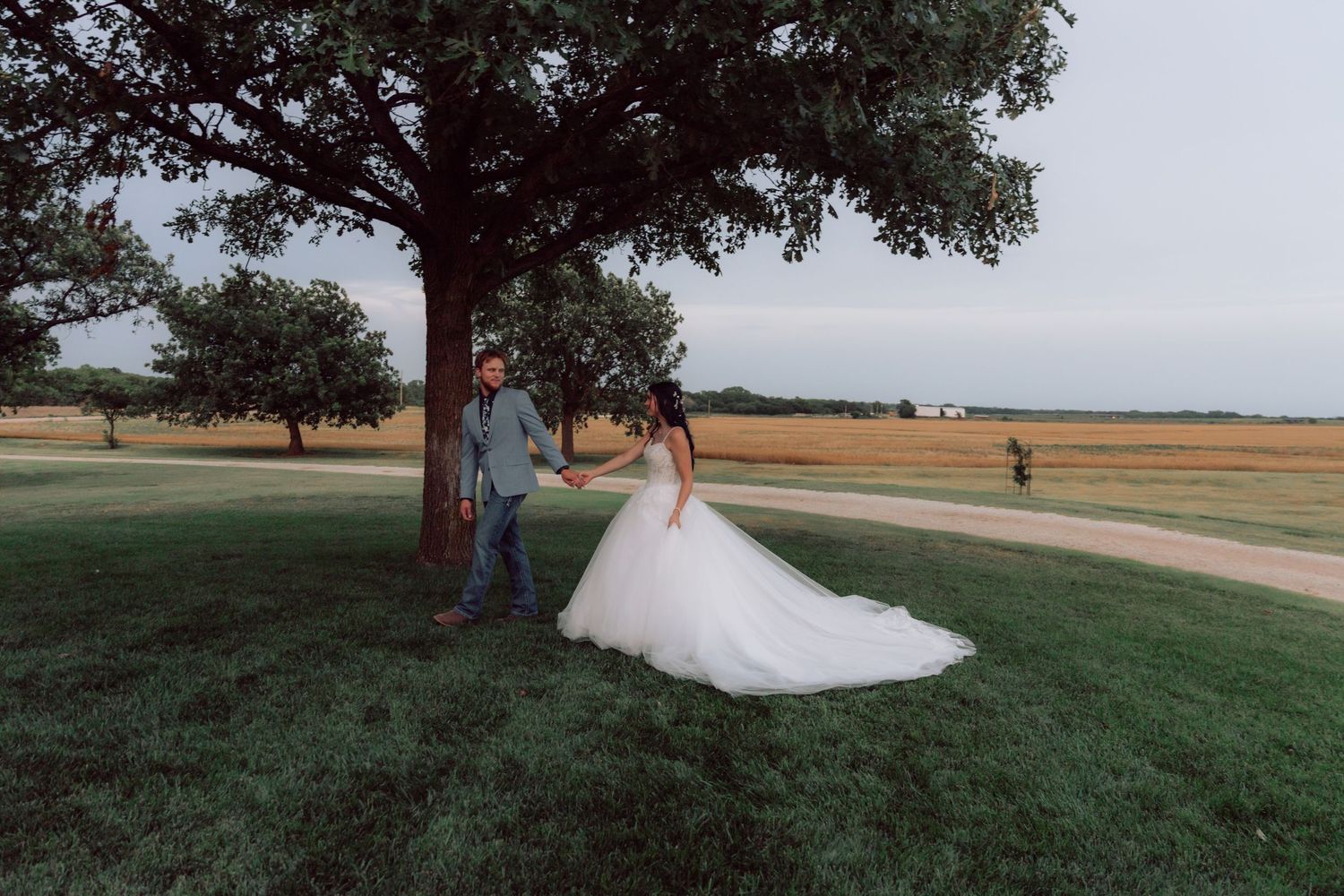Wedding couple poses beneath a large tree in a serene countryside setting.