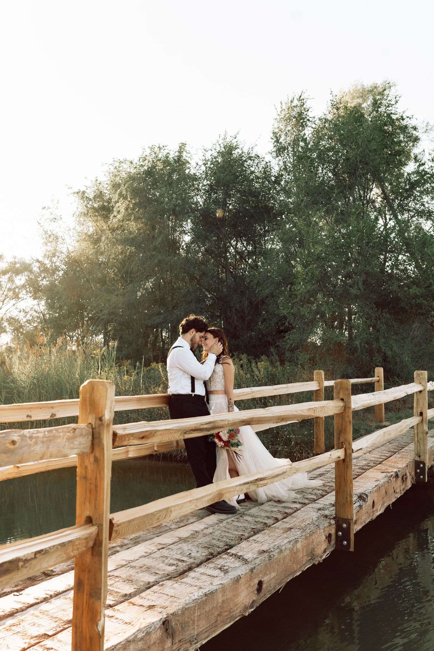 Newlyweds embrace on weathered wooden bridge during golden hour.