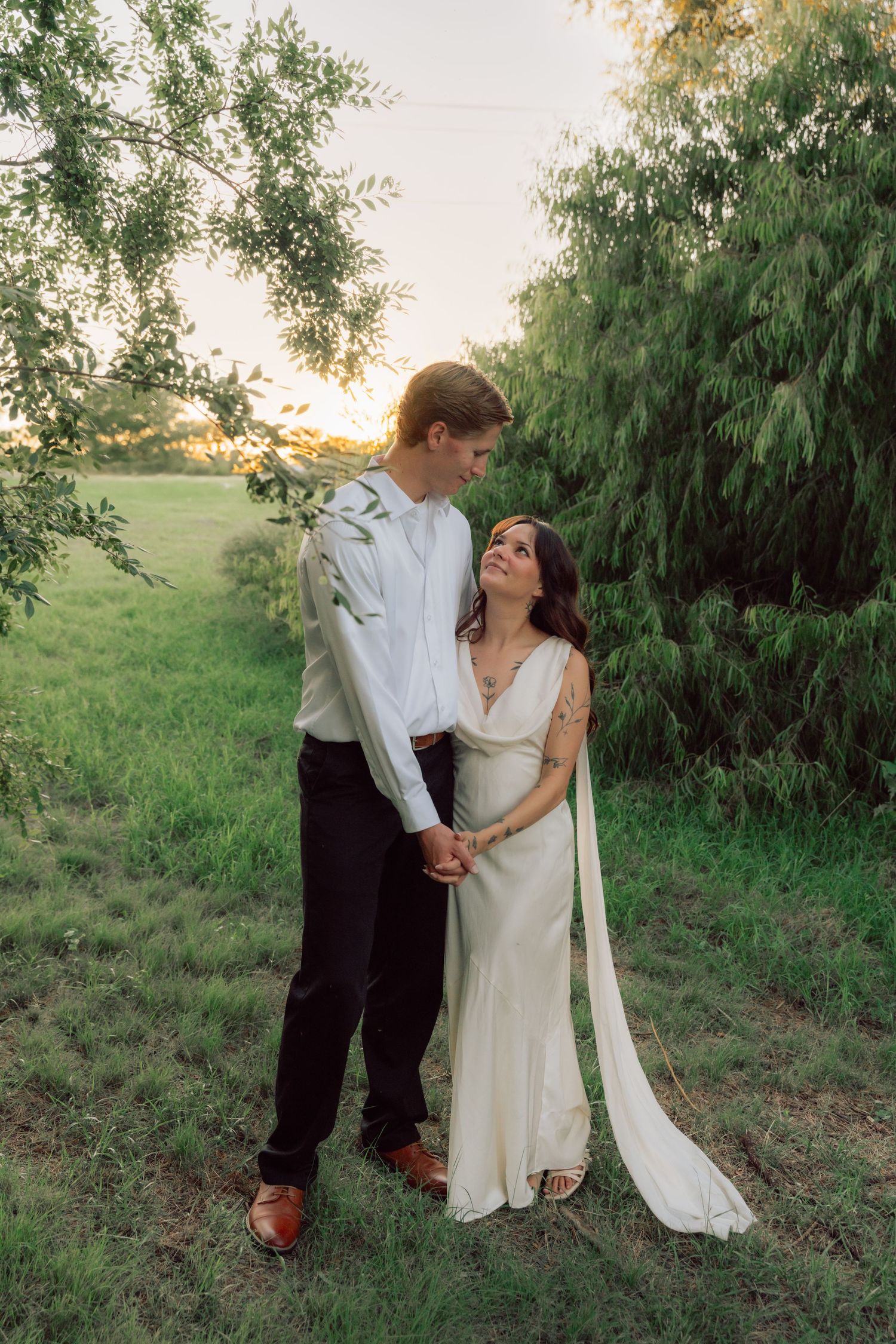 A couple in wedding attire shares an intimate moment beneath willow trees at sunset.