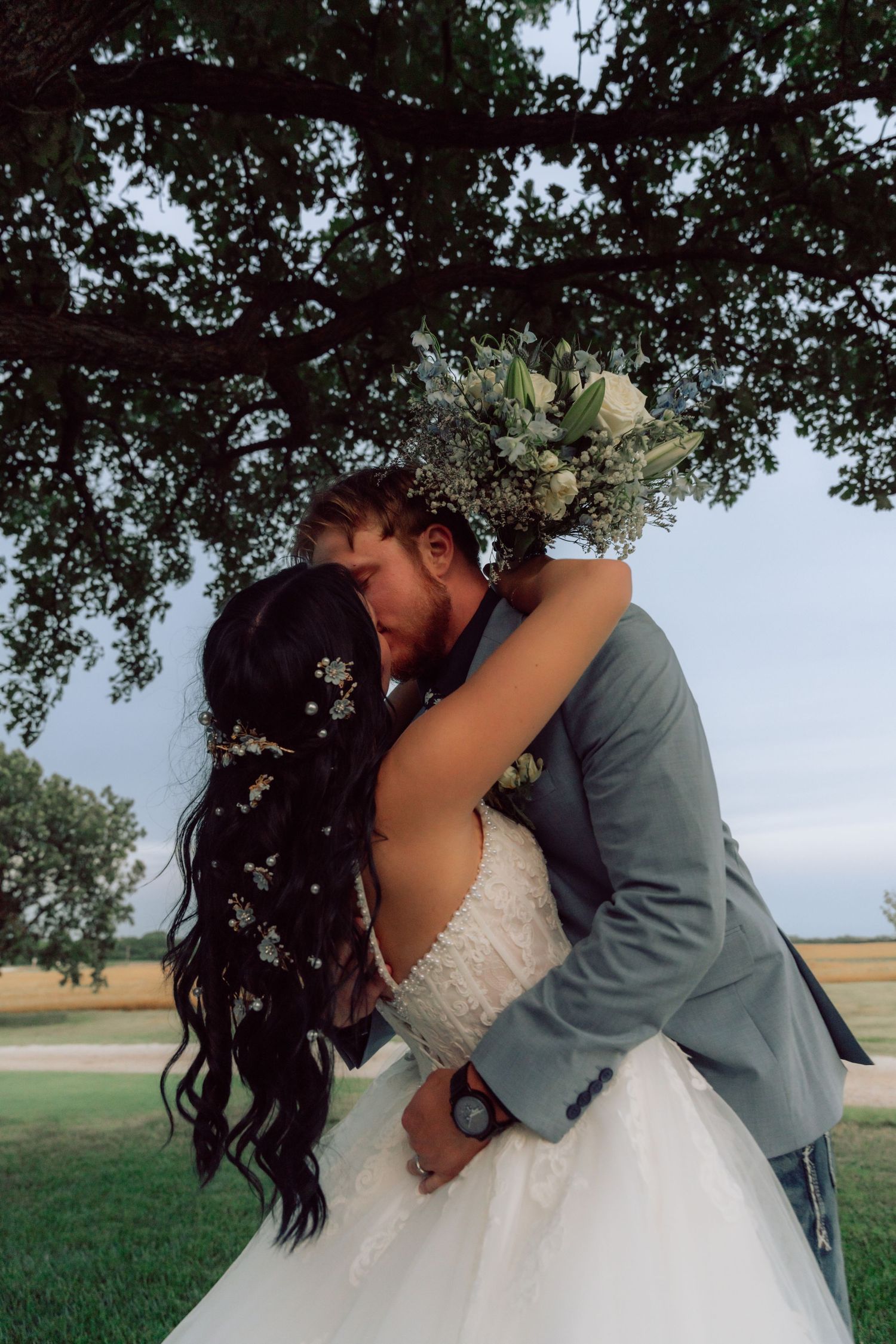 Romantic outdoor wedding portrait with couple kissing under overhanging tree branches.