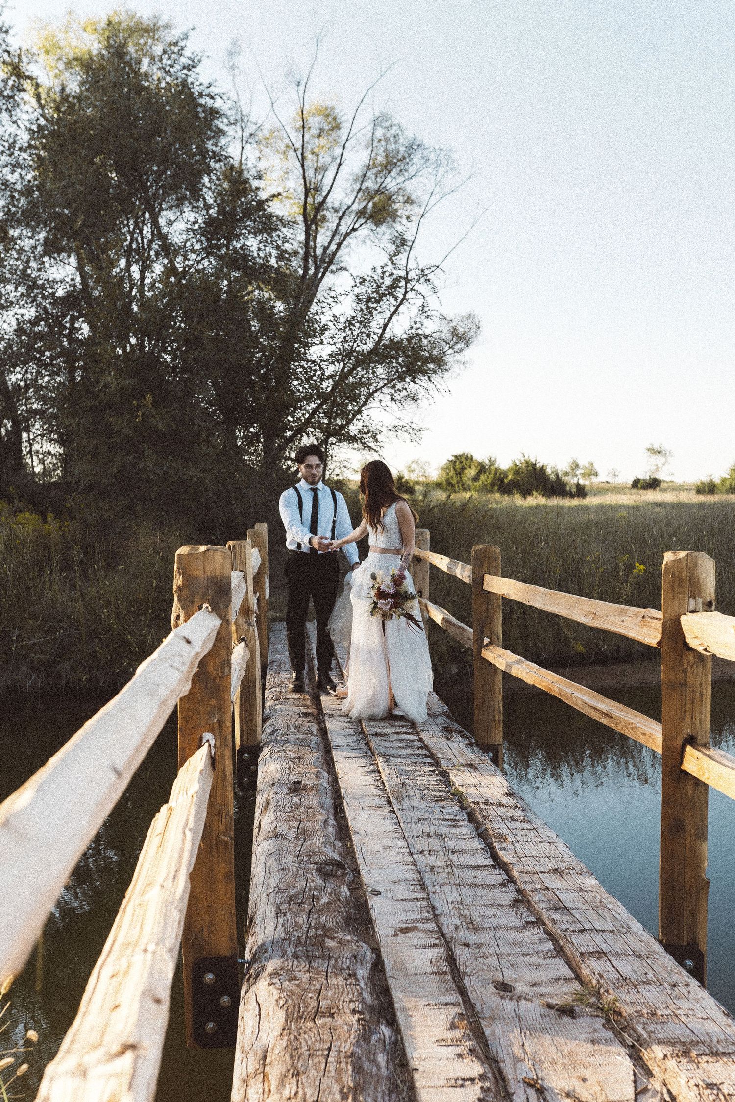 Couple in wedding attire poses on rustic wooden bridge over peaceful water.