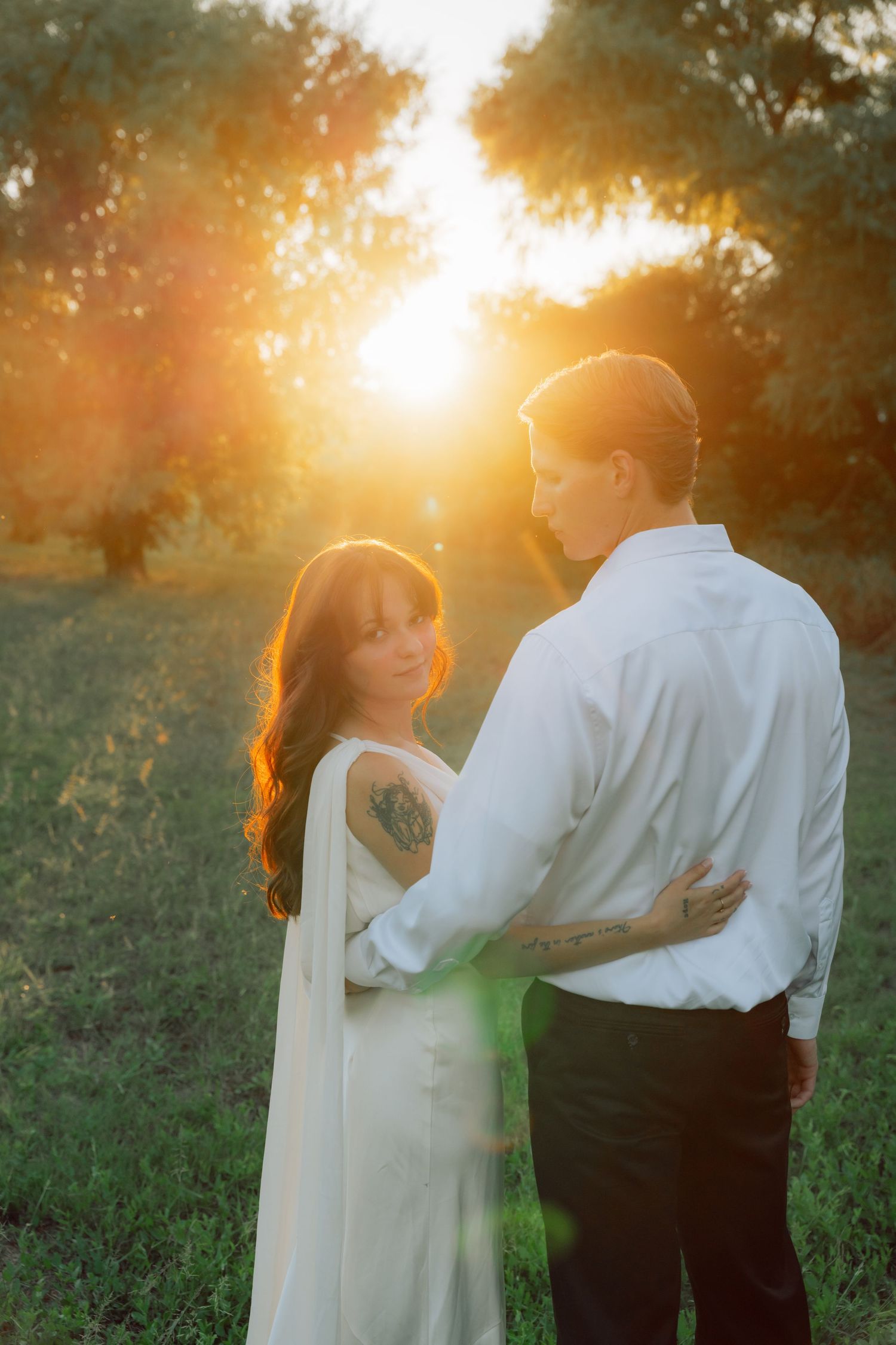 A tender embrace between two people in white and black attire against a sunset-lit field.