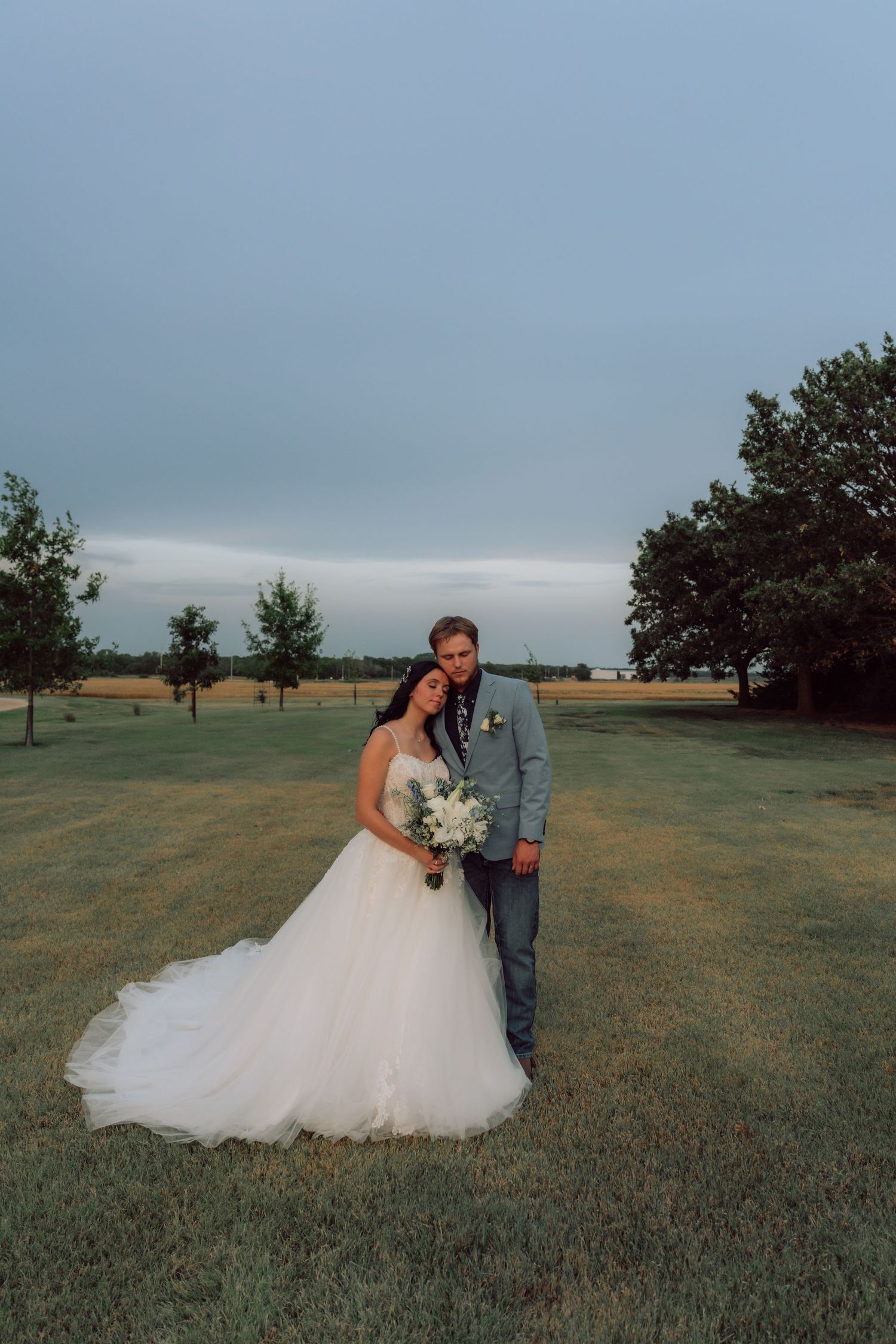 Wedding couple poses for romantic portraits in an open field with overcast sky.