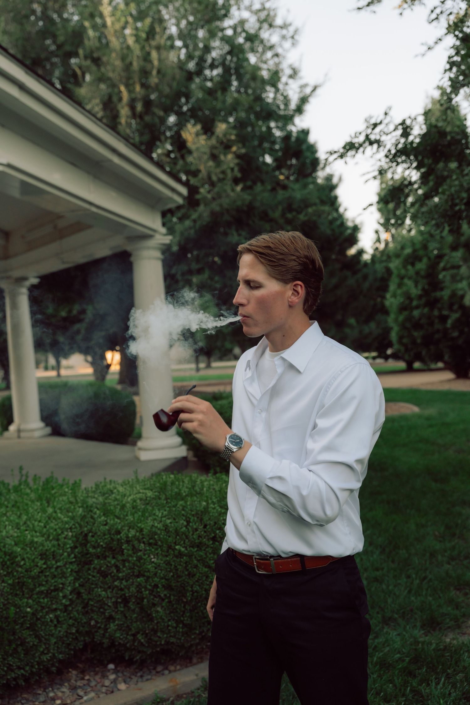 A person in white shirt and dark pants stands near white columns in a garden setting.