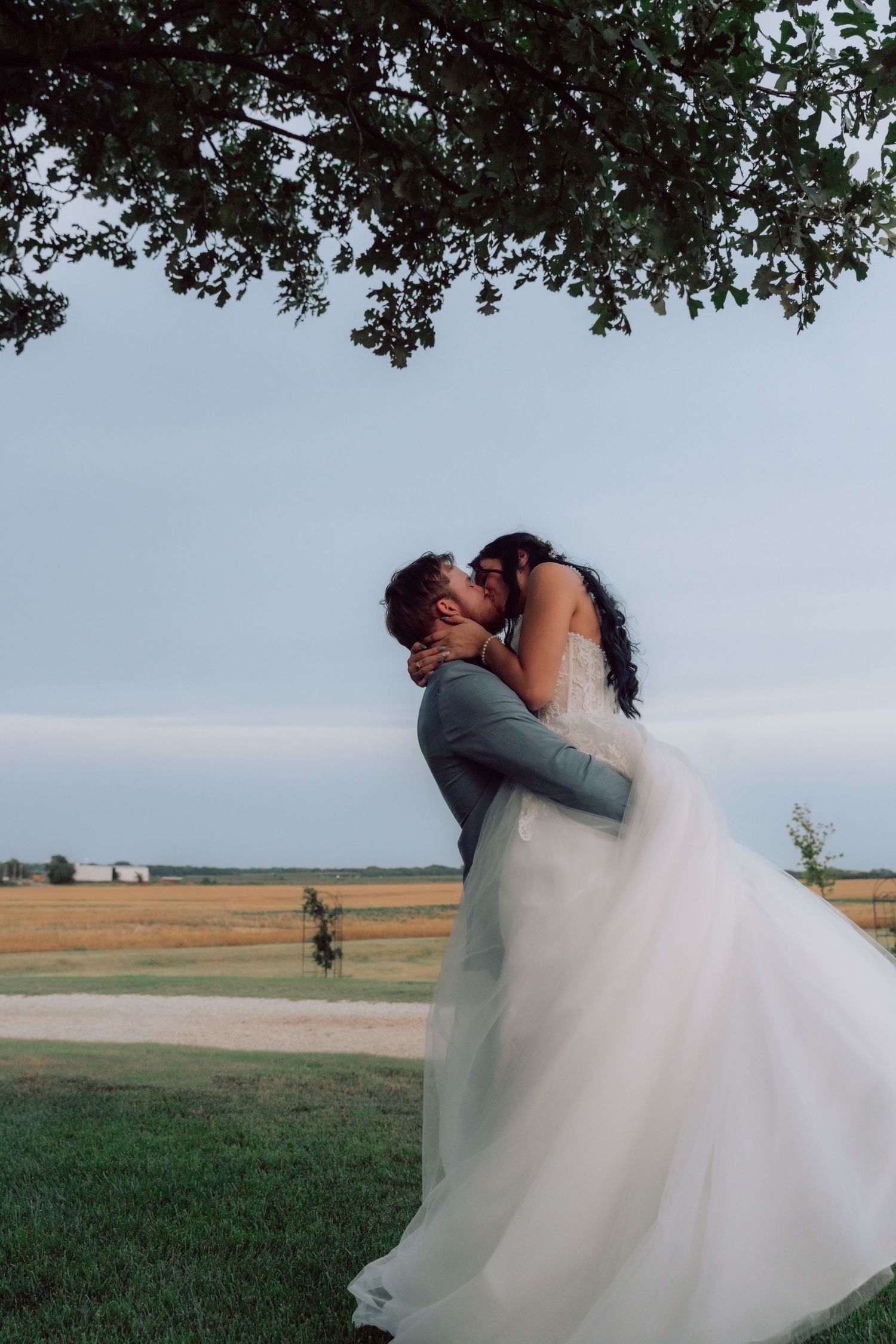 A bride and groom share a romantic dip kiss against a rural landscape backdrop.