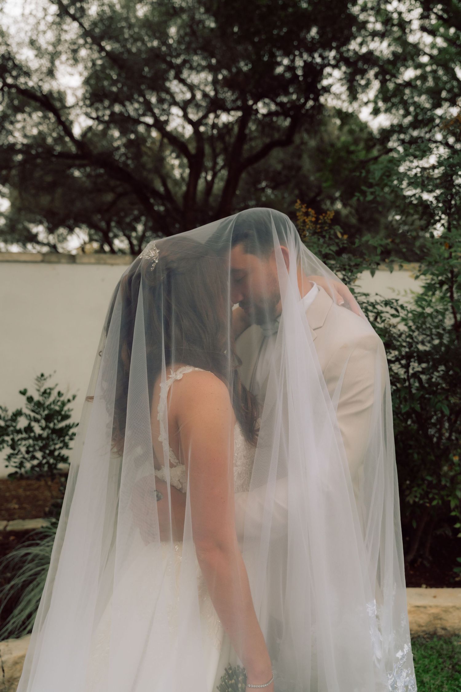 A tender moment shared under a flowing white wedding veil in a garden setting.