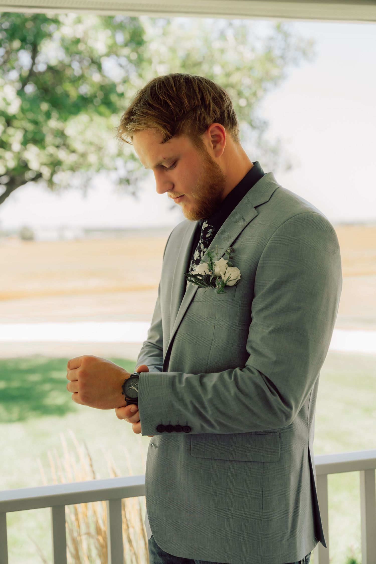 A person in a grey suit adjusts their boutonniere while standing on a porch.