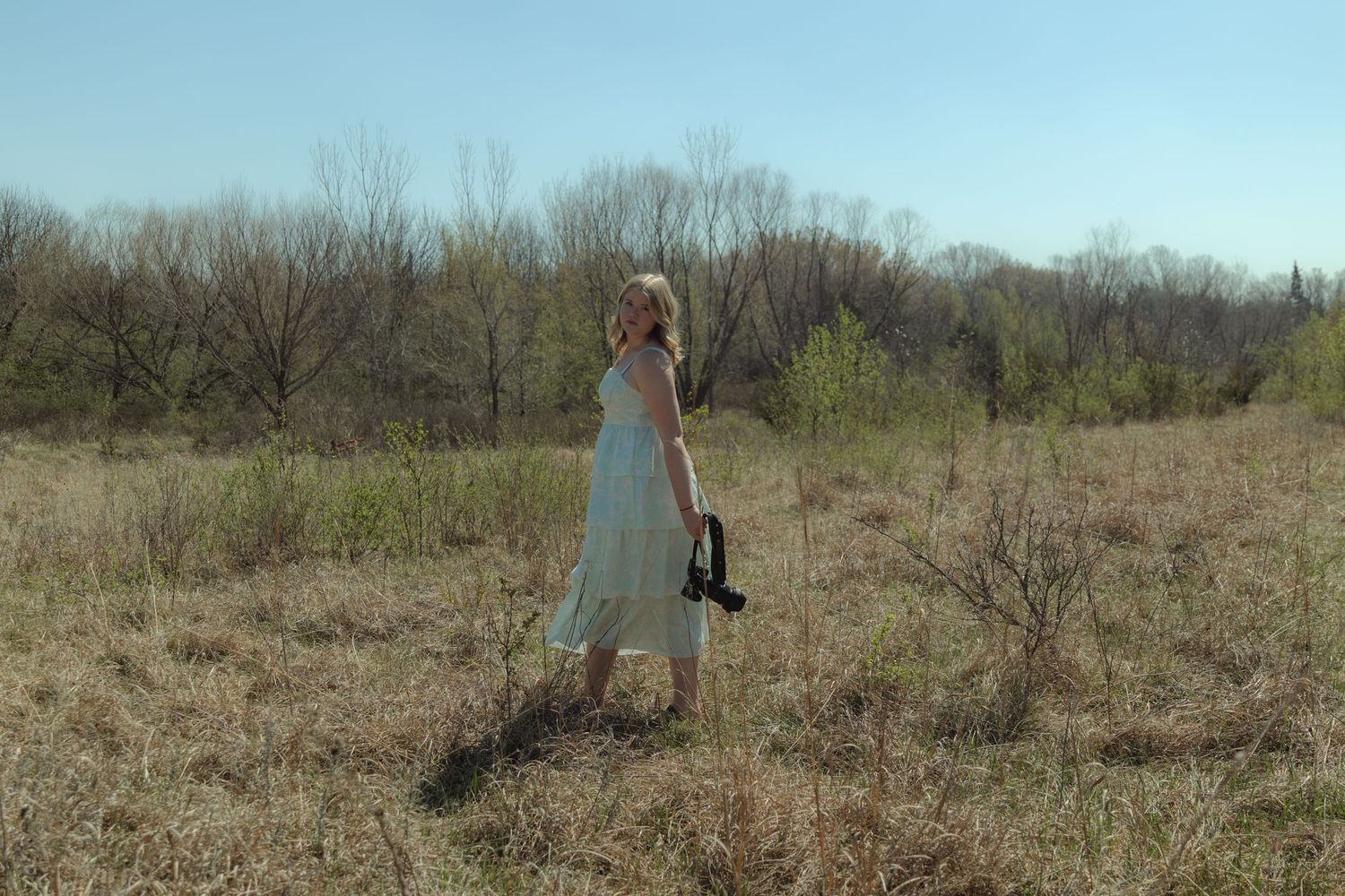 Woman in white dress holding shoes standing in a rustic field with bare trees and blue sky.