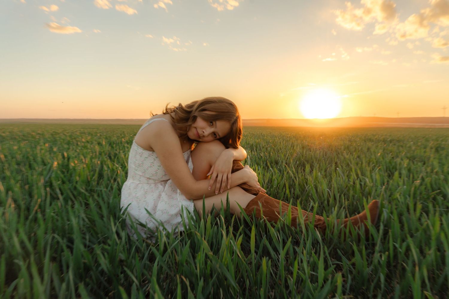 Woman in white dress sitting in green field at golden sunset, hugging knees peacefully.