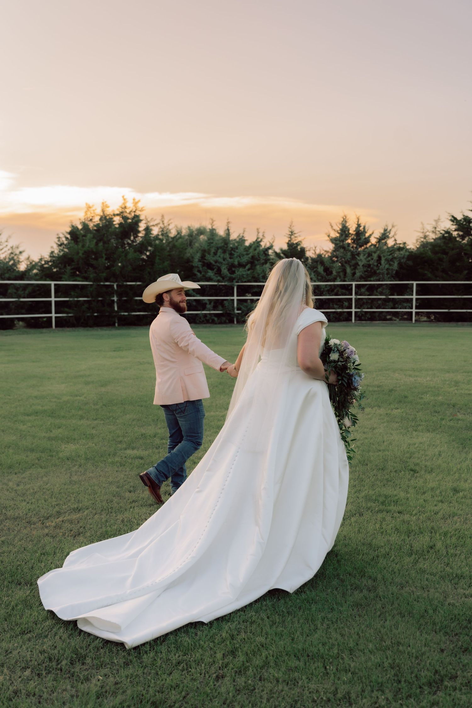 A bride and groom walk hand in hand across a grassy field at sunset.