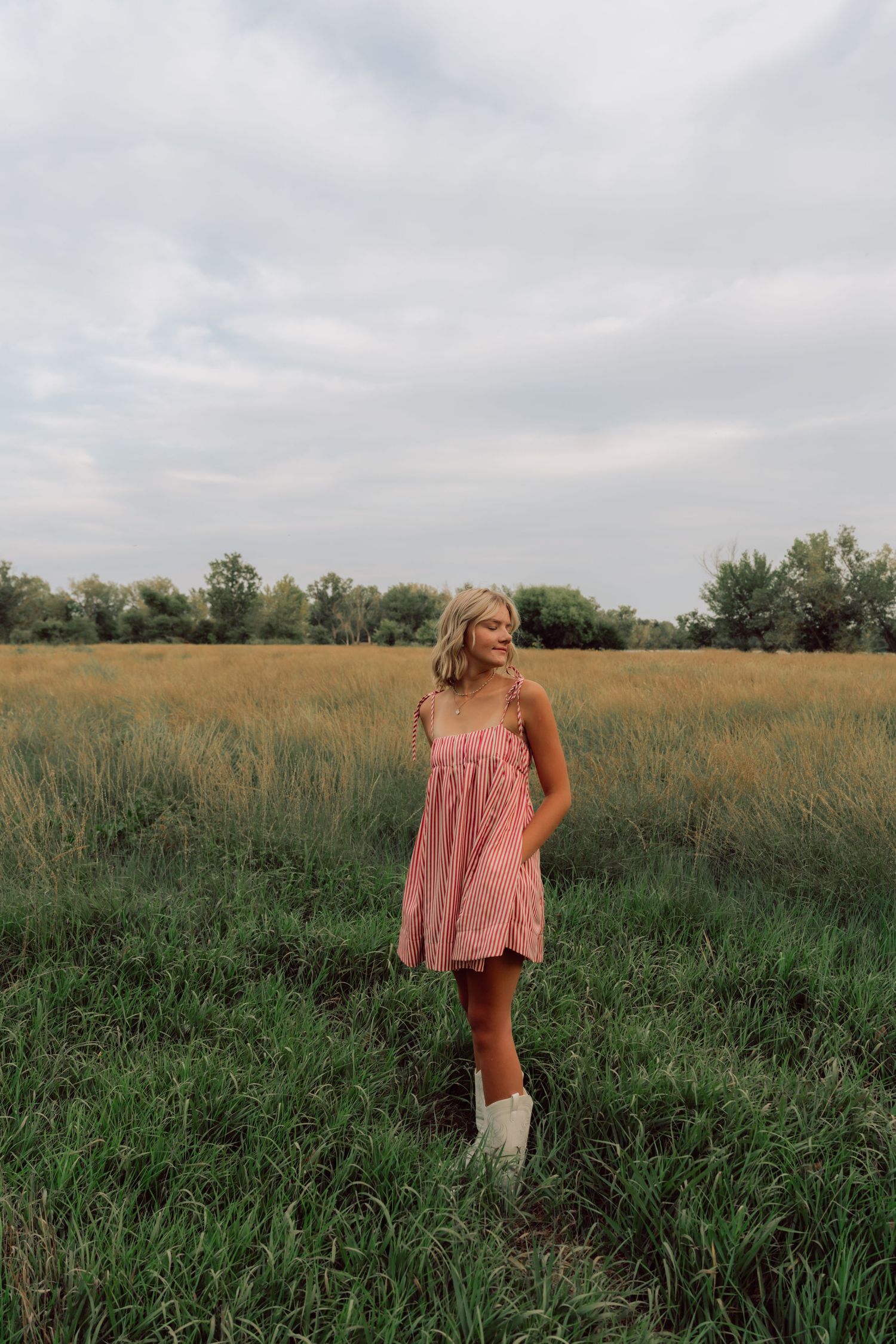 Figure wearing a flowy pink dress stands gracefully in tall grass under cloudy skies.
