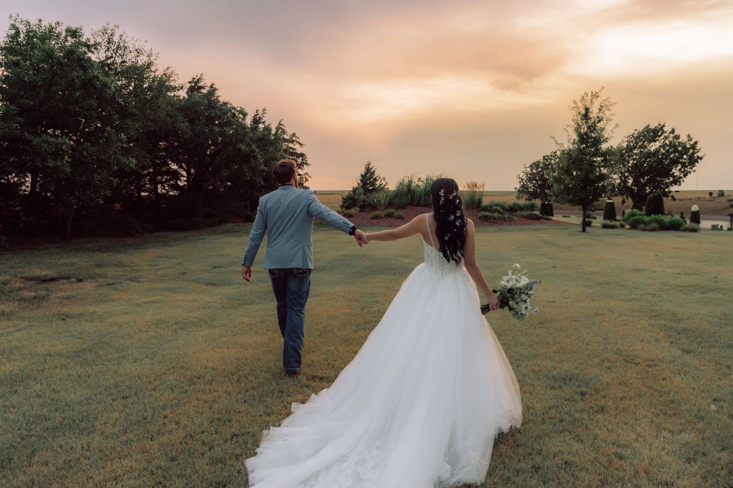 A wedding couple walks hand in hand across a sunset field with the bride's dress trailing behind.