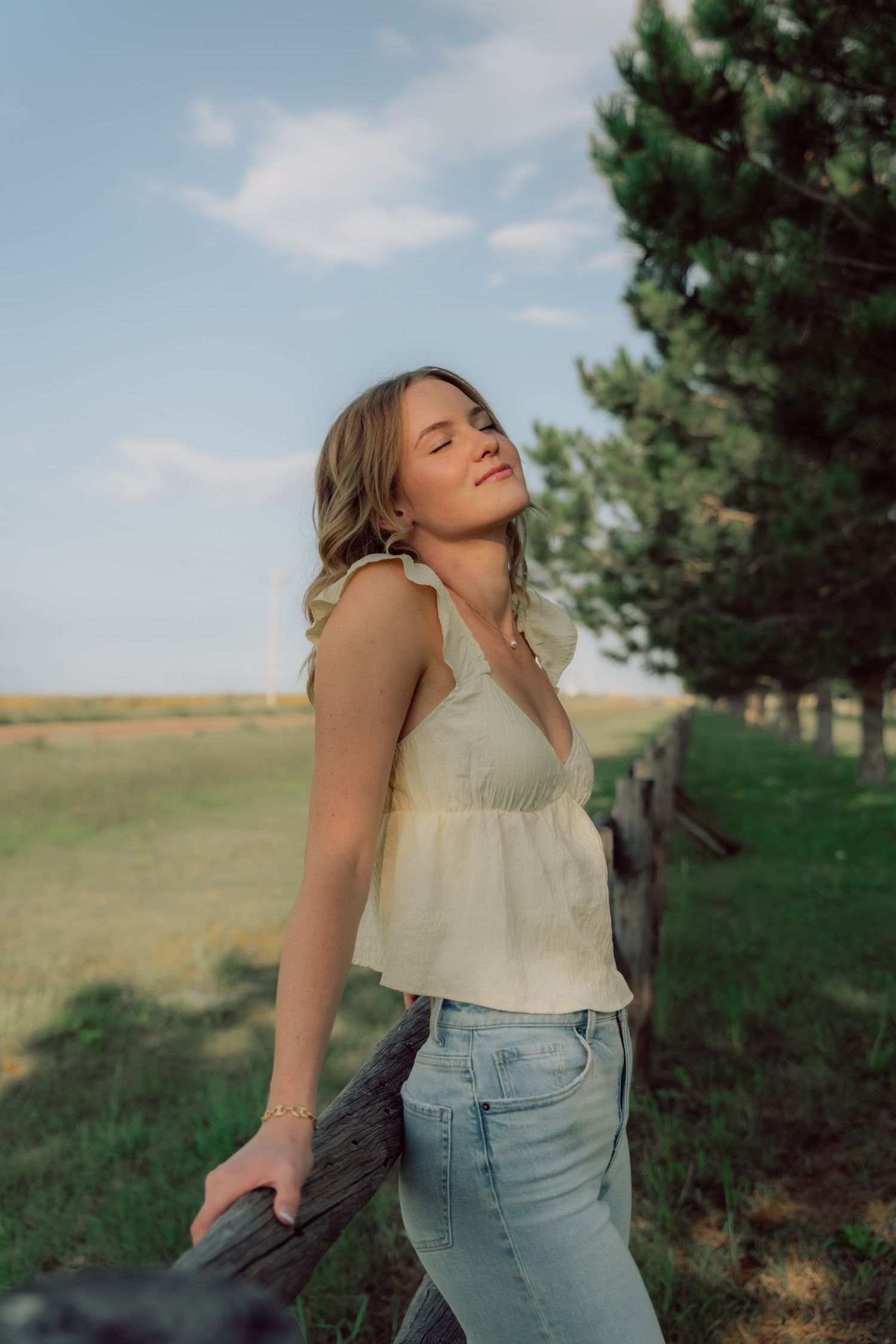 A woman in a cream top and jeans poses casually against a rural backdrop with pine trees.