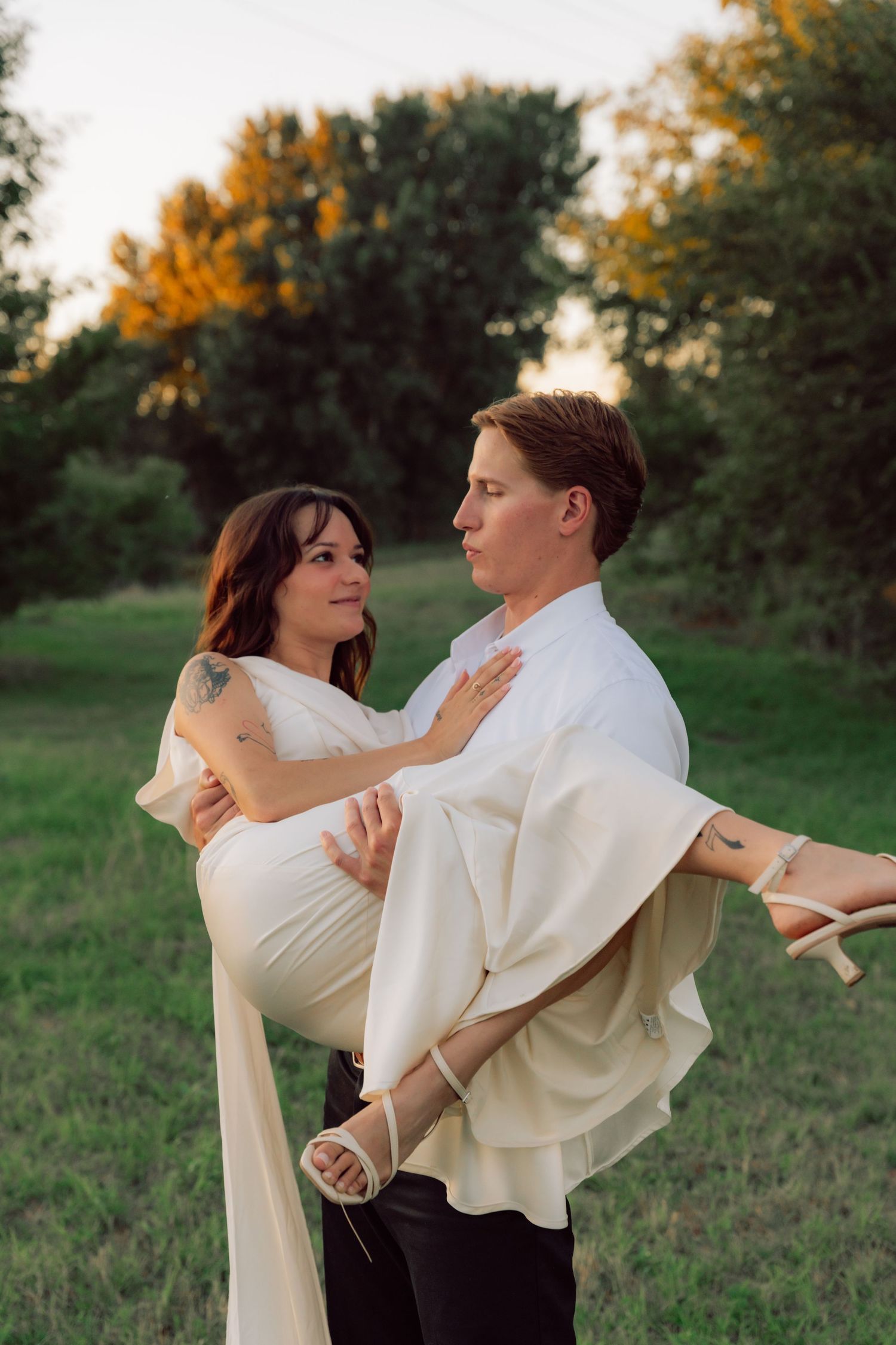 A groom in white carries his bride in an elegant white dress during their outdoor photoshoot.