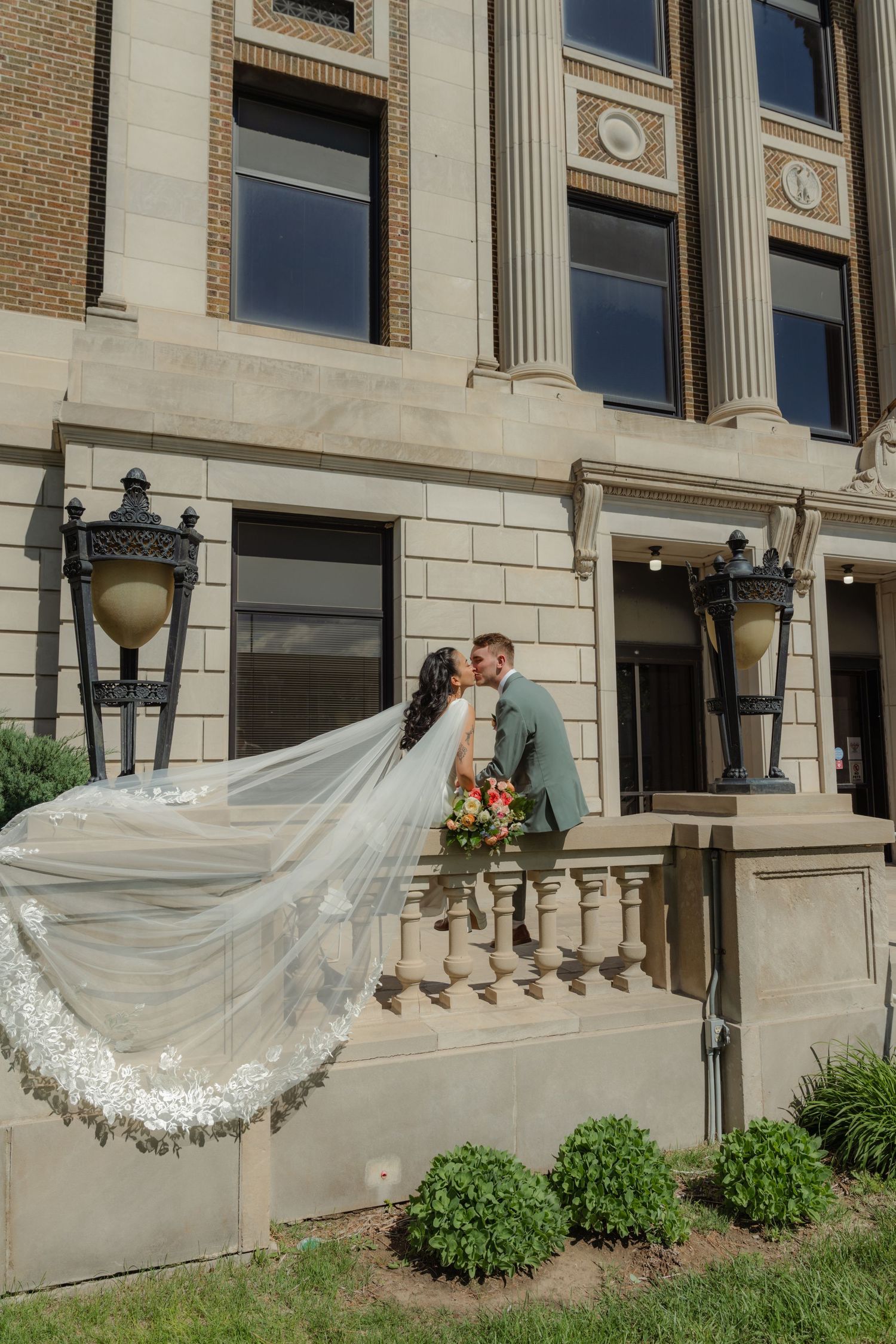 Bride and groom kissing on elegant stone steps with flowing cathedral veil, holding bouquet outside classic building.