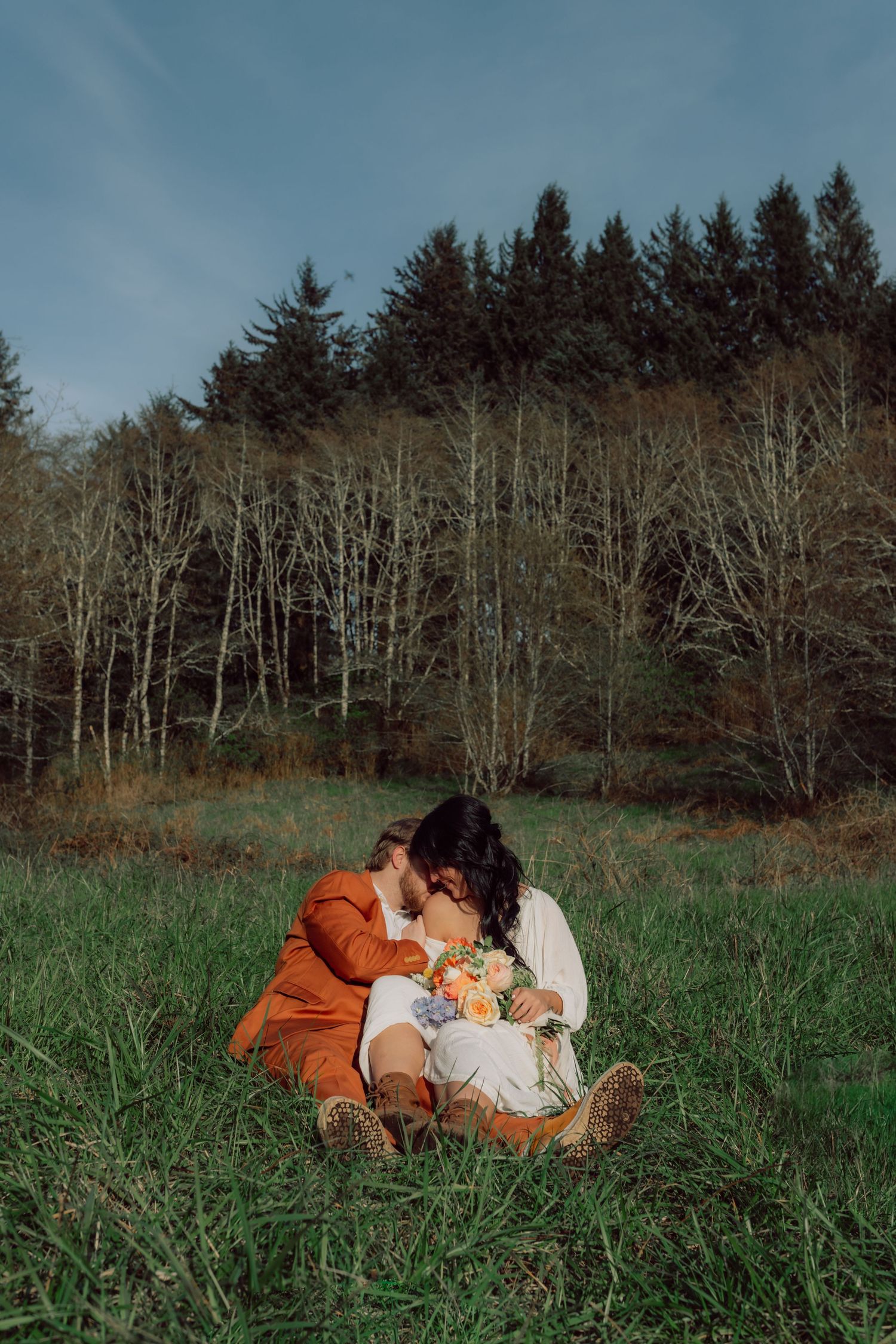 Couple embraces on orange blanket in green field with forest backdrop.