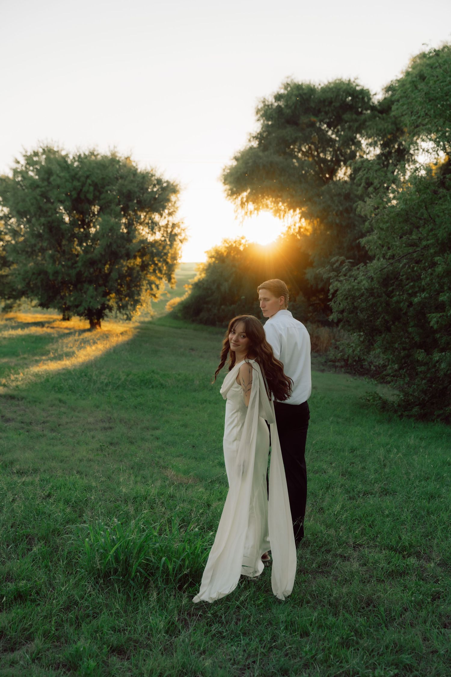 A romantic couple embraces in a sunlit meadow during golden hour with trees in the background.