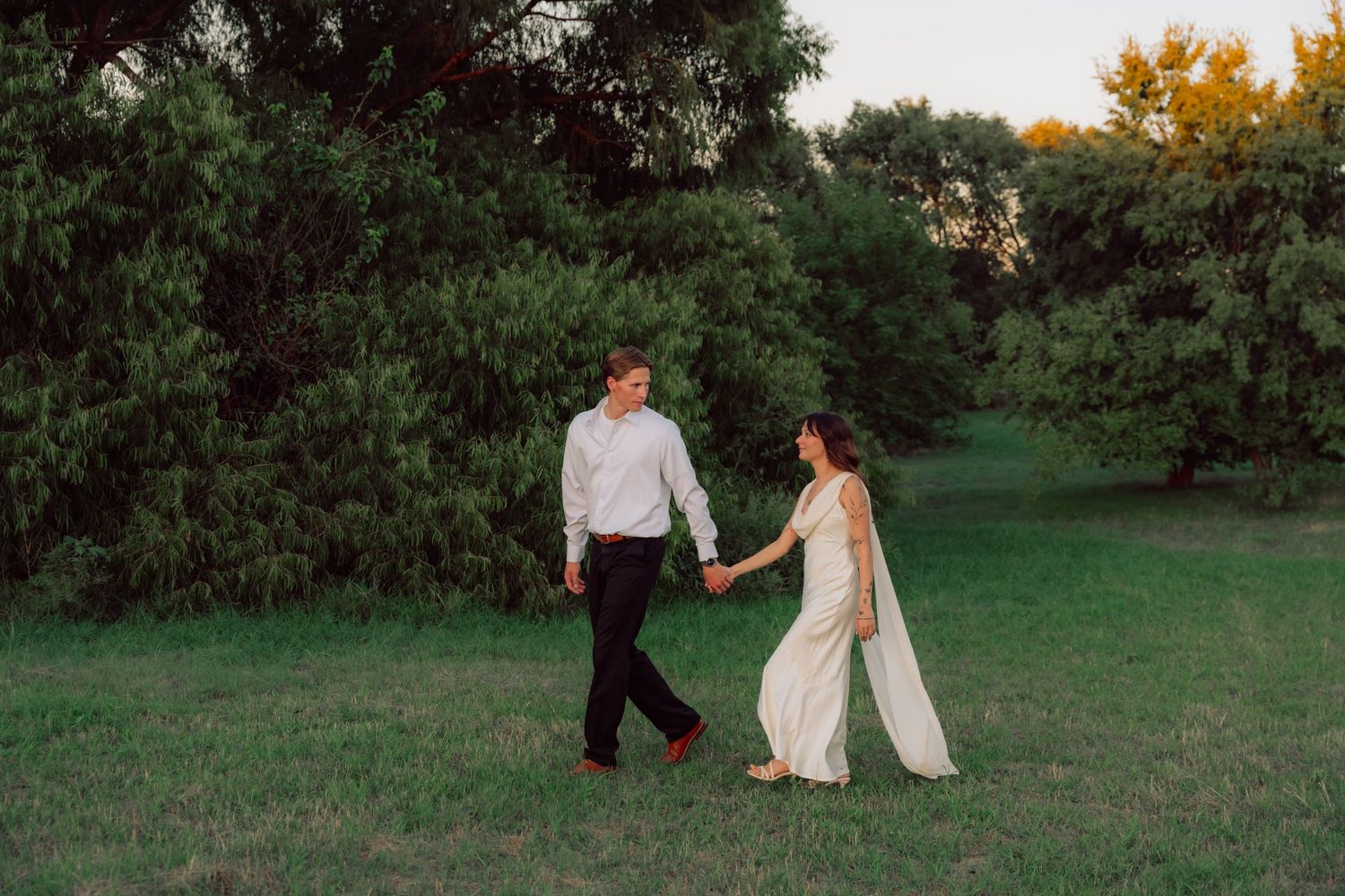 A couple in wedding attire walks hand in hand through a lush green field at dusk.