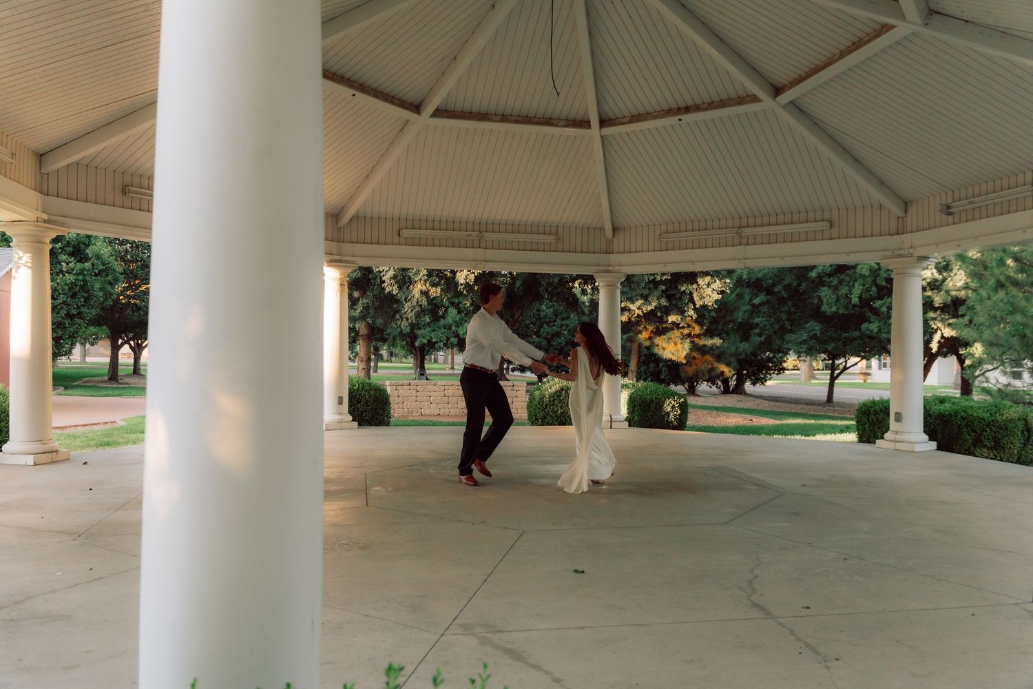 A couple dances together under a white octagonal gazebo in a park setting.