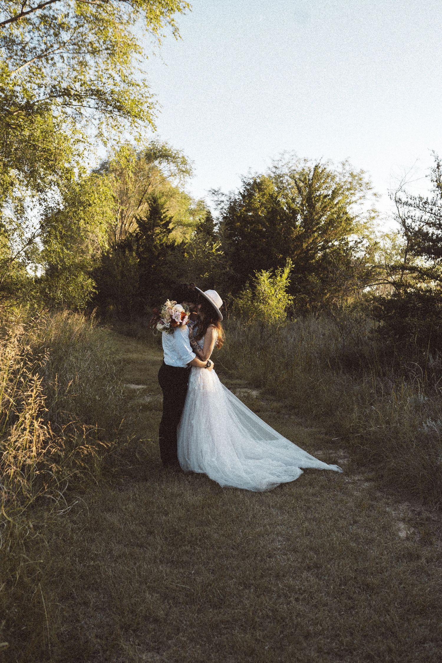 A romantic wedding couple embraces on a grassy hillside path during golden hour.