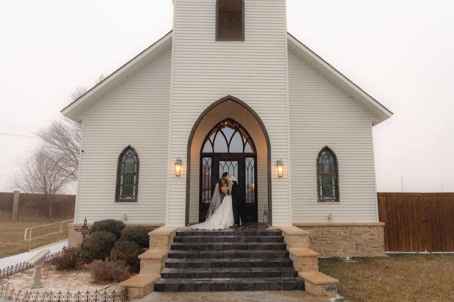 Small white country church with arched wooden doors and stained glass windows sits in a rural setting on a cloudy day.