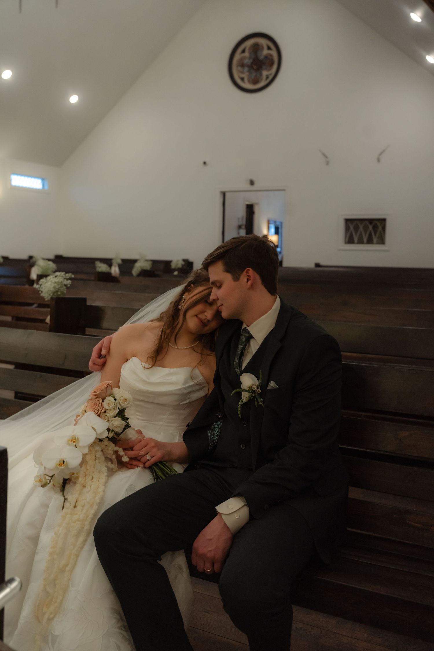 Newlywed couple sitting close together on church pew, bride in white gown with bouquet, groom in dark suit.