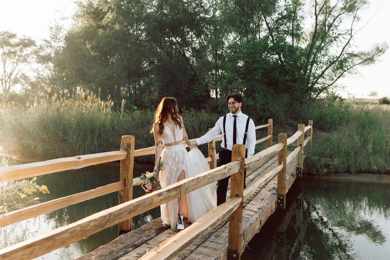 Two people stroll hand in hand across a weathered wooden footbridge surrounded by nature.