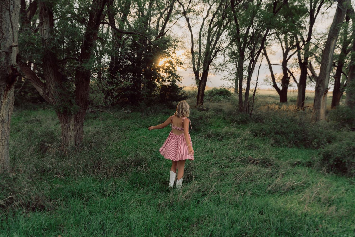 Figure in pink dress walking through wooded area at dusk.