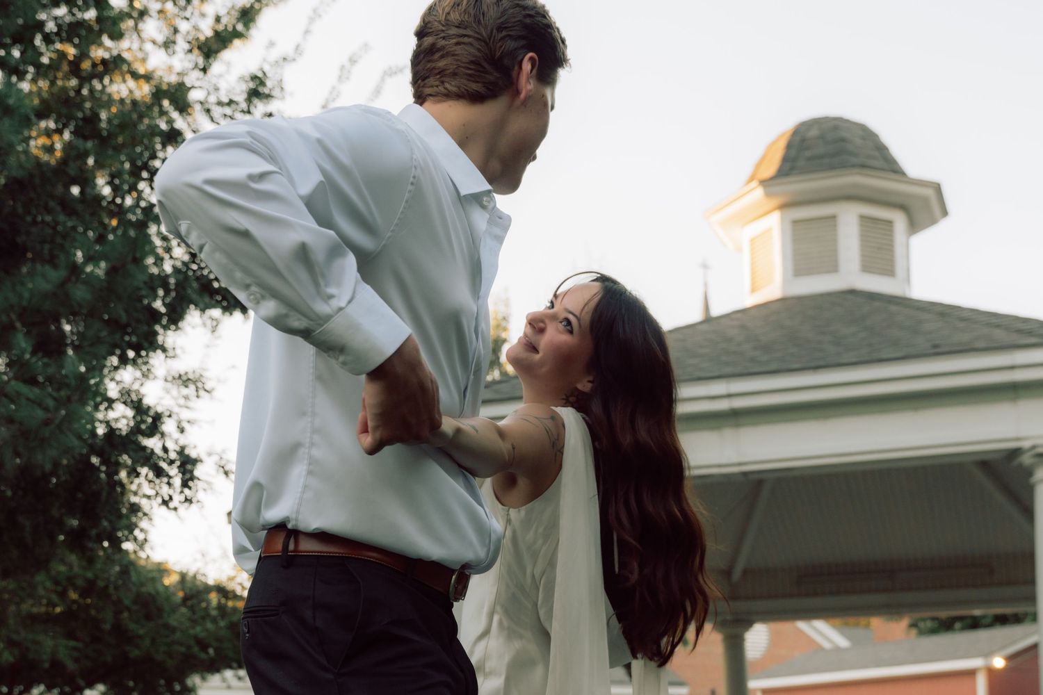 A couple embraces near white columns and greenery during golden hour.